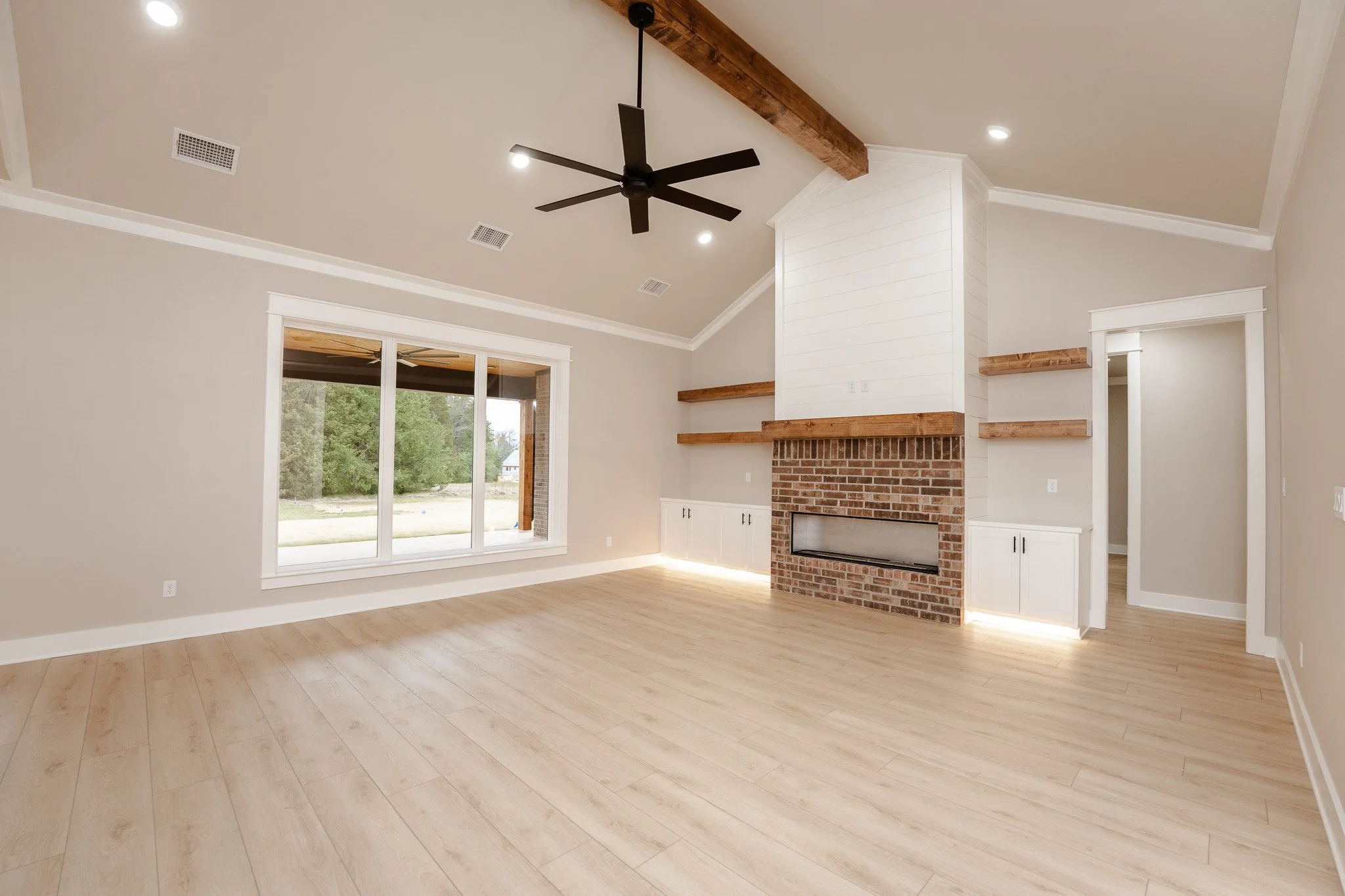 Empty living room with large window, brick fireplace, beige walls, light wood flooring, and ceiling fan.