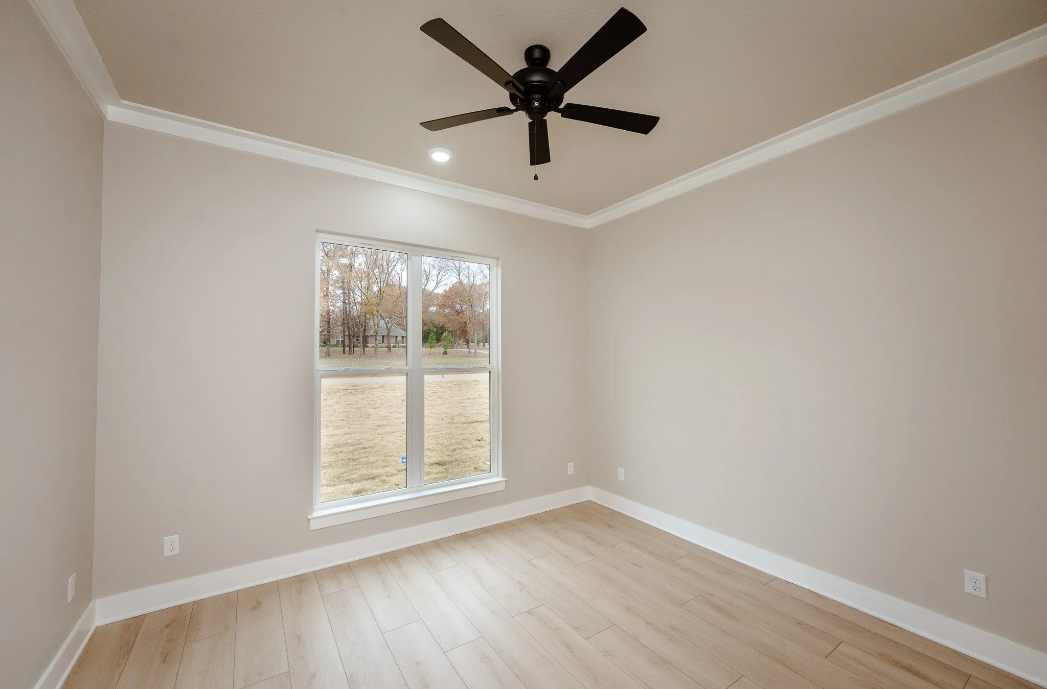 Empty room with beige walls, a single window, a black ceiling fan, and light wood flooring.