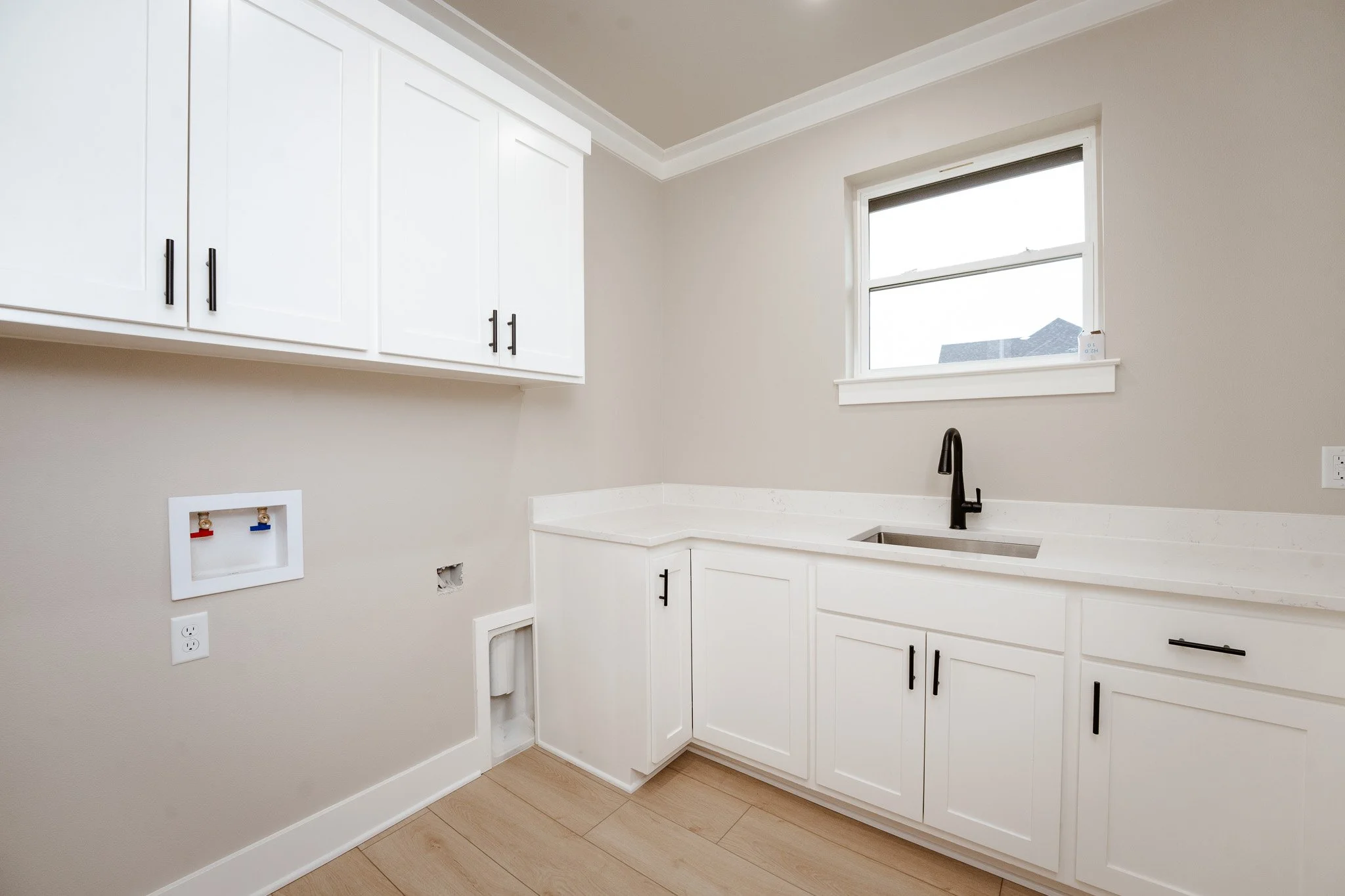 Empty laundry room with white cabinets, a black faucet, a window, and laundry hookups on beige walls.