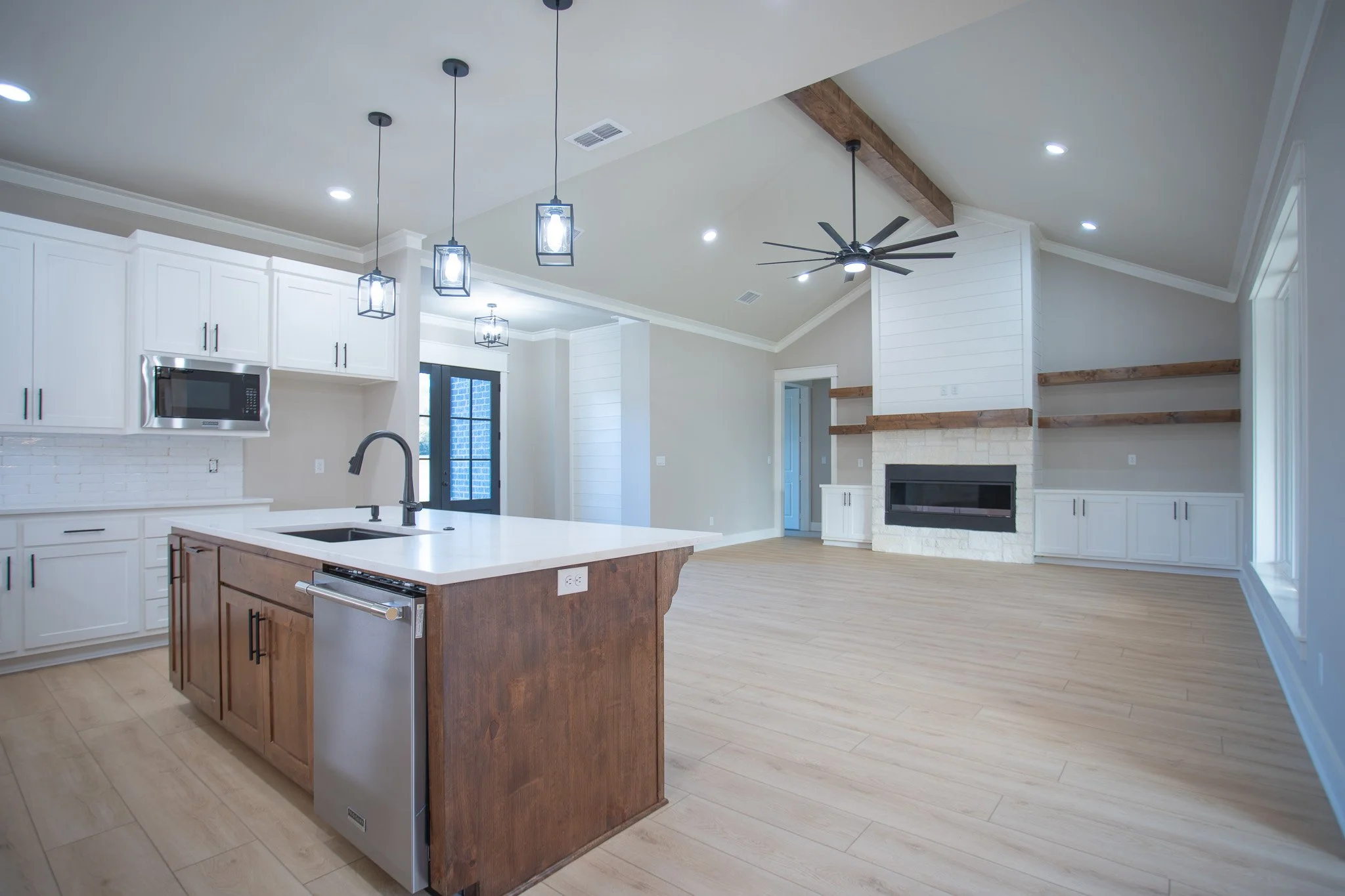 Open-concept living room and kitchen with white cabinets, a kitchen island, and a white brick fireplace with wooden shelves.