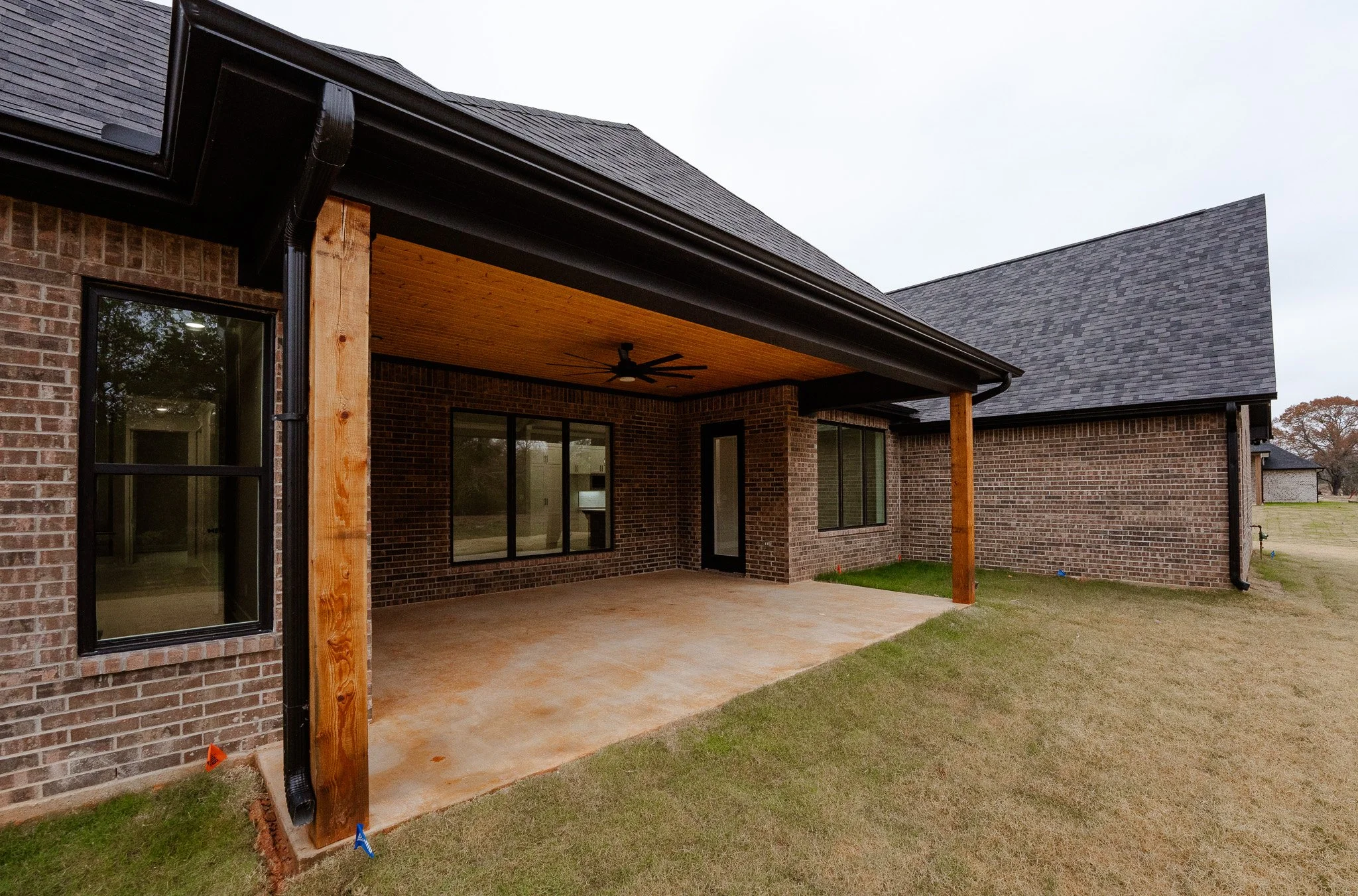 Backyard porch of a brick house with a wooden ceiling, ceiling fan, sliding glass doors, and windows, with a grassy lawn and neighboring houses in the background.