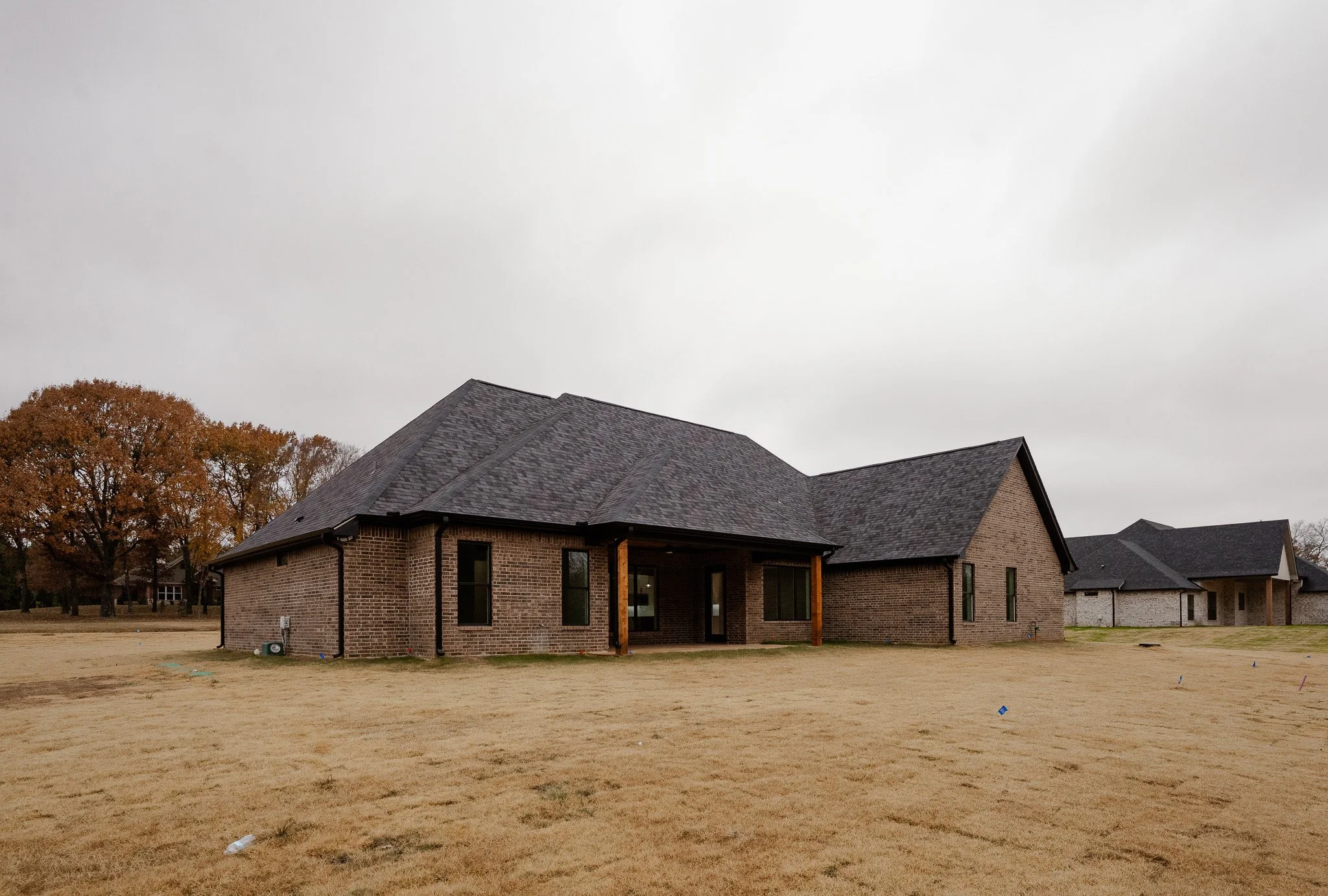A brick house with a dark shingled roof, small covered patio, and a yard with dry grass under an overcast sky.