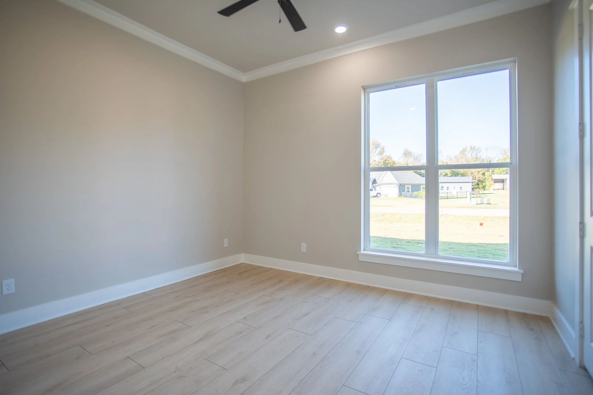 Empty room with large window, light-colored wooden floor, beige walls, white baseboards, ceiling fan, and recessed lighting.