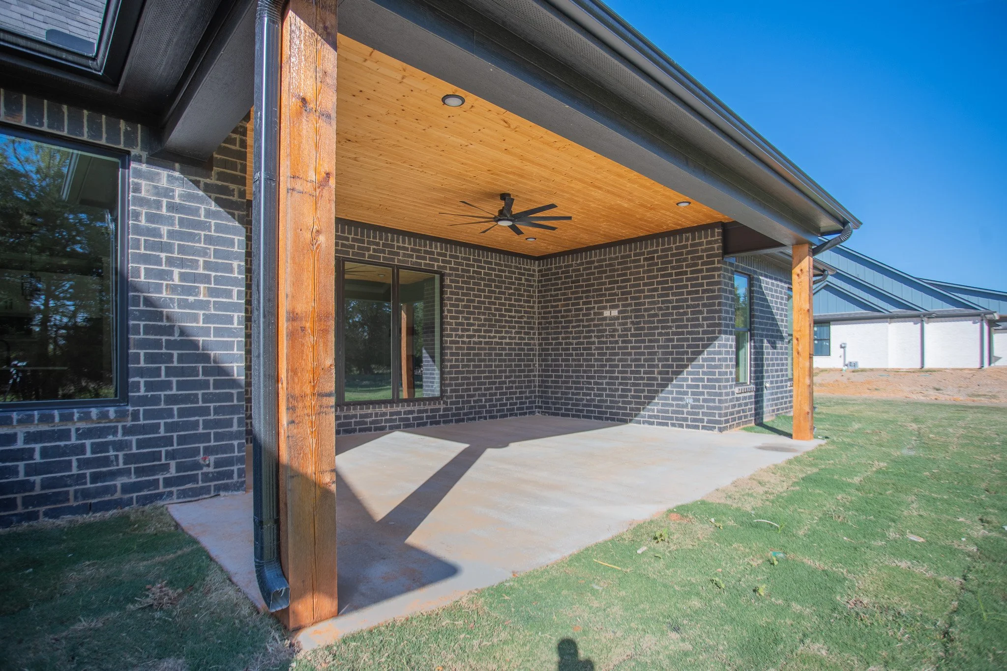 Newly constructed house with black brick walls, a wooden ceiling on the porch, a ceiling fan, and an outdoor lawn area.