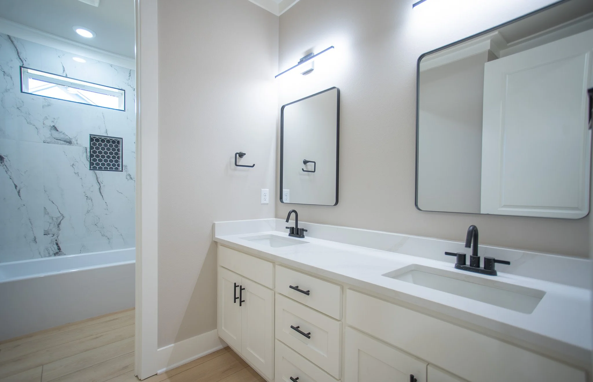 Modern bathroom with double vanity, white cabinetry, black fixtures, and a marble shower visible in the background.