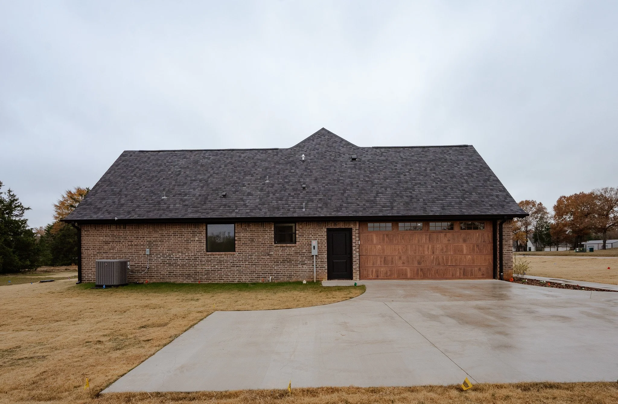 Front view of a brick house with a black door and a wooden garage door on a concrete driveway, with grass on either side, under a cloudy sky.