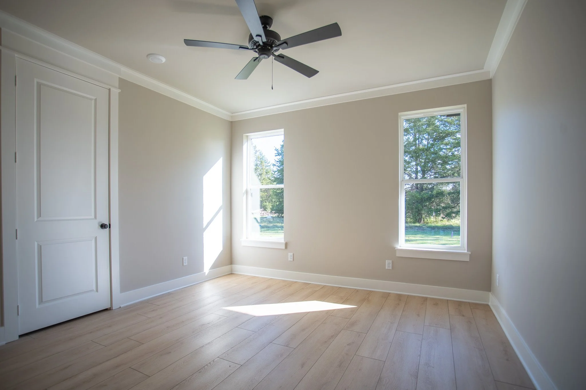 Empty room with two windows, a ceiling fan, beige walls, and light wood flooring.