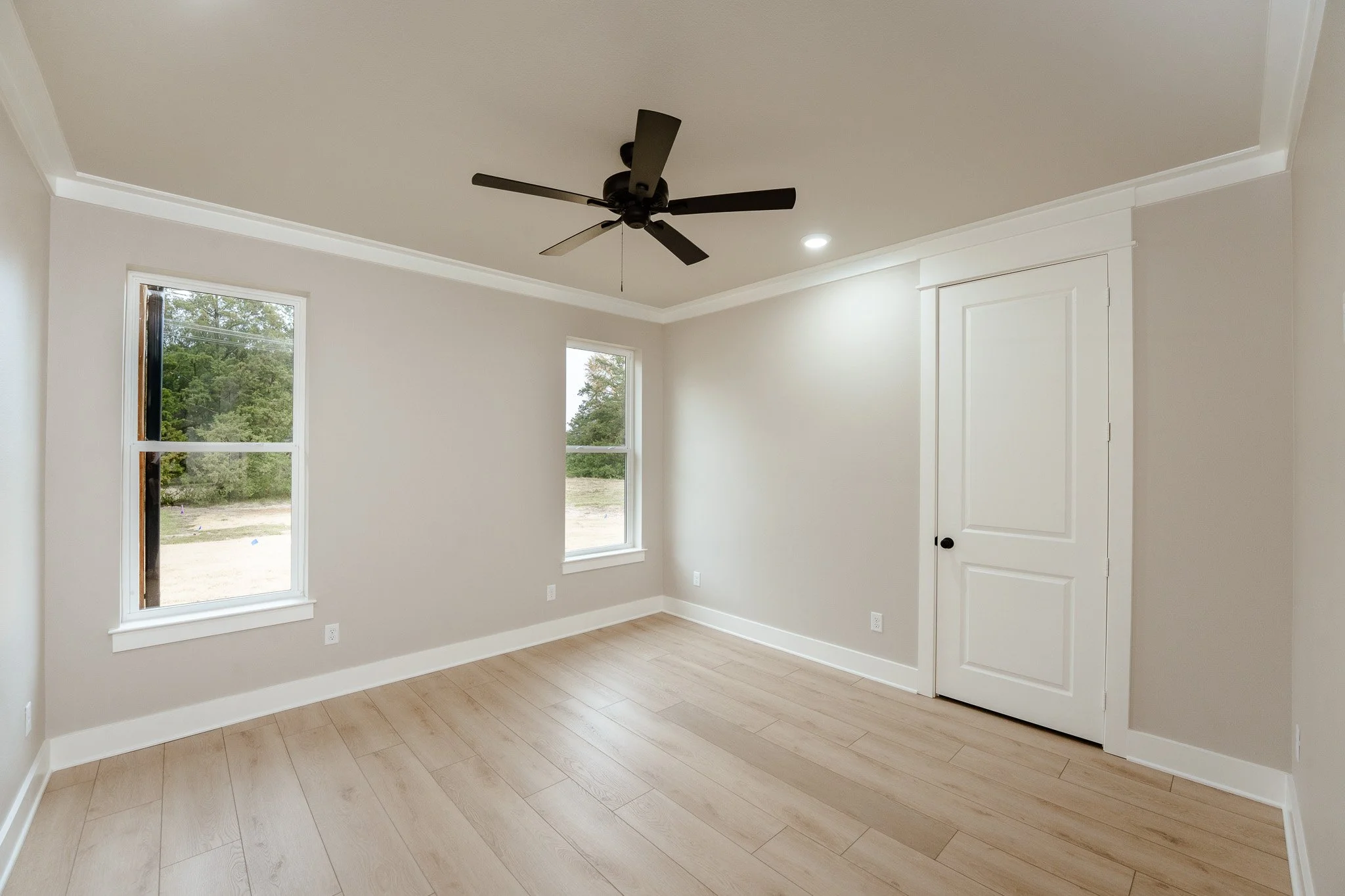 Empty bright room with large windows, white walls, ceiling fan, and light wood flooring.