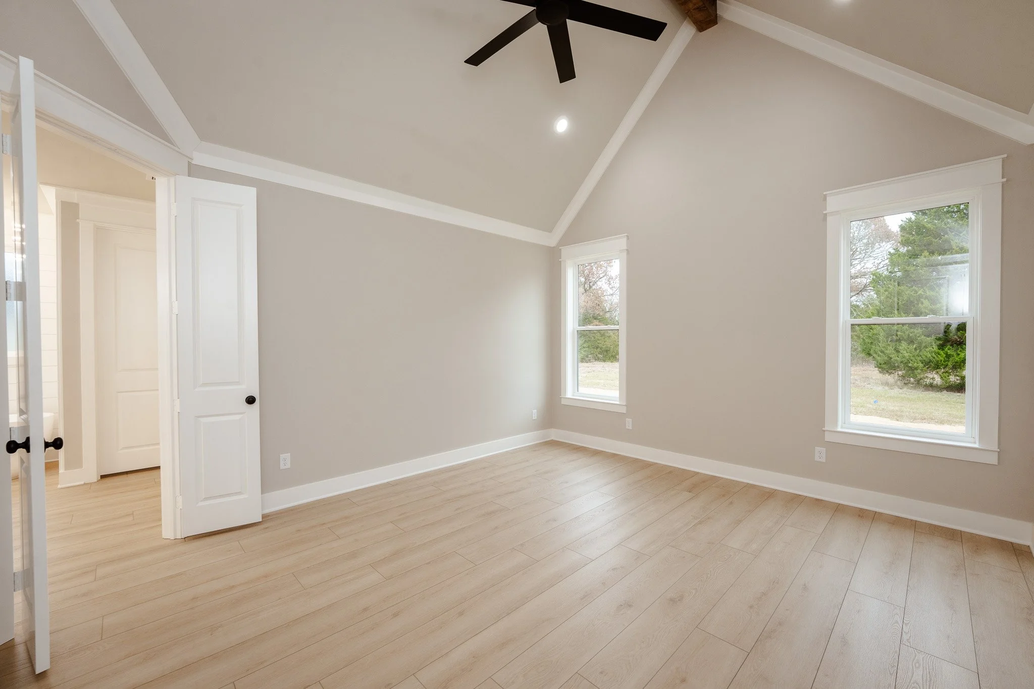 Empty room with light wood flooring, beige walls, white trim, two large windows showing trees outside, an open white door, and a black ceiling fan.