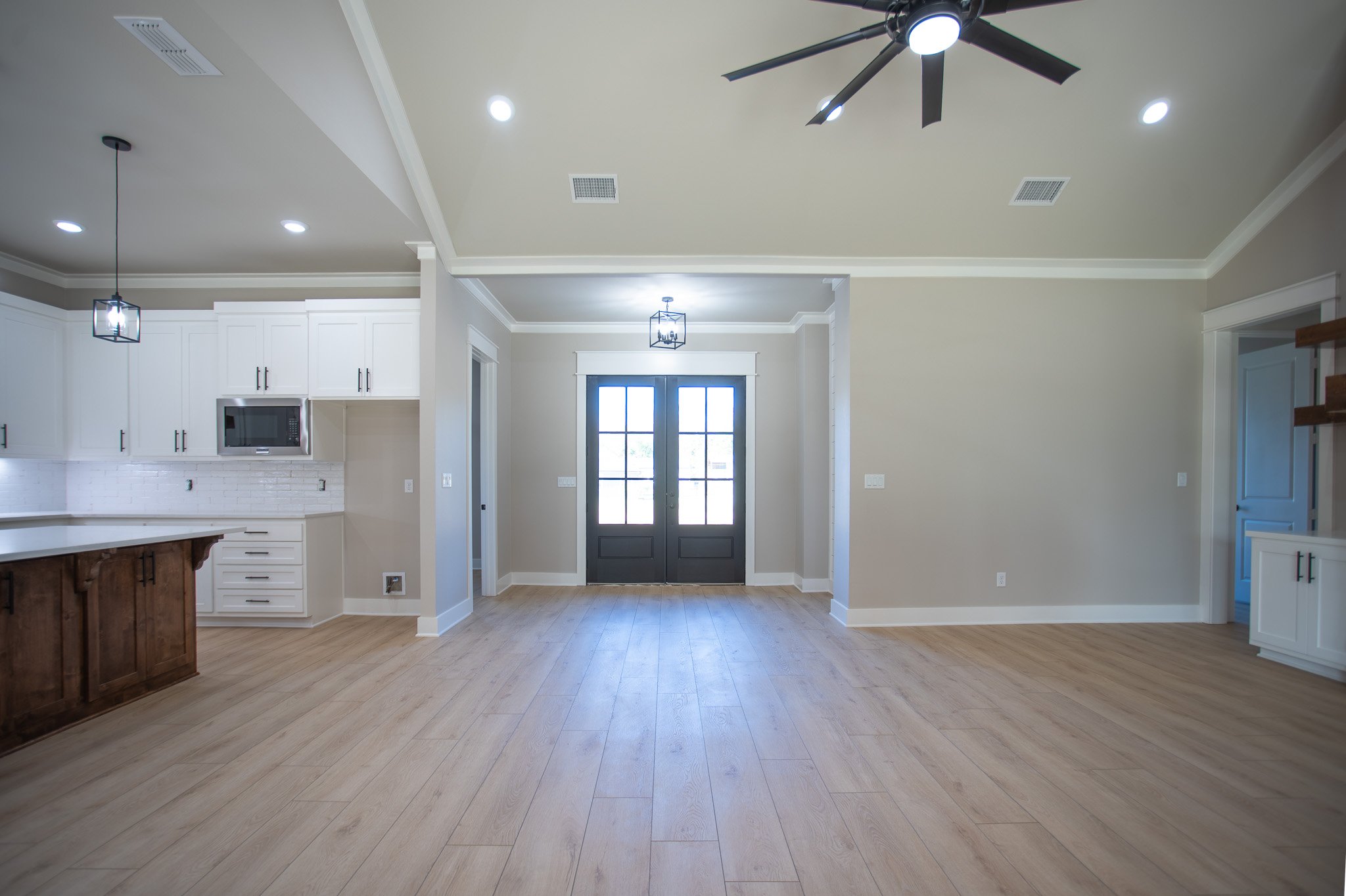 Empty kitchen and dining area with light wood flooring, white cabinets, a black front door, and modern lighting fixtures.