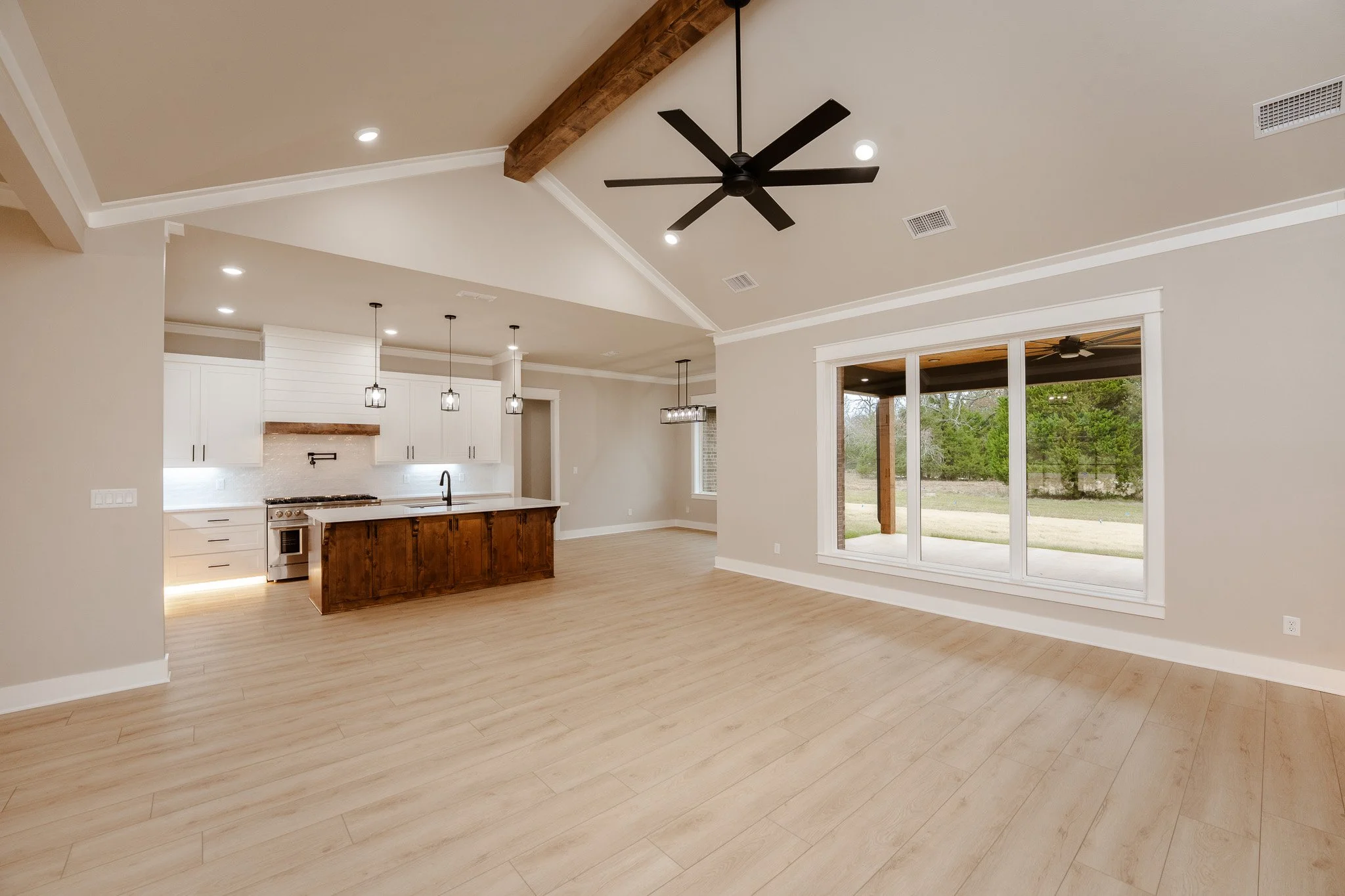 Empty living room and kitchen with large window, wooden floor, ceiling fan, and modern light fixtures.