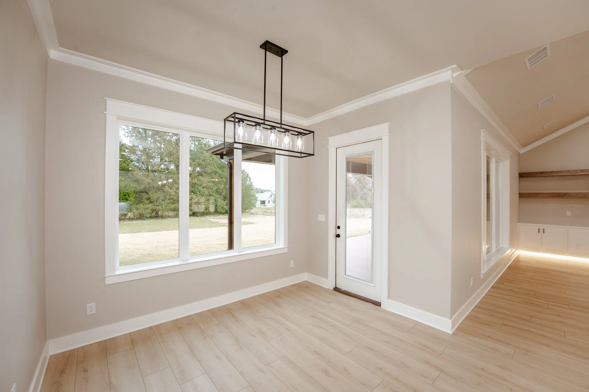 Empty room with large window and glass door, wooden flooring, modern black chandelier, neutral-colored walls, and built-in shelves with lighting.