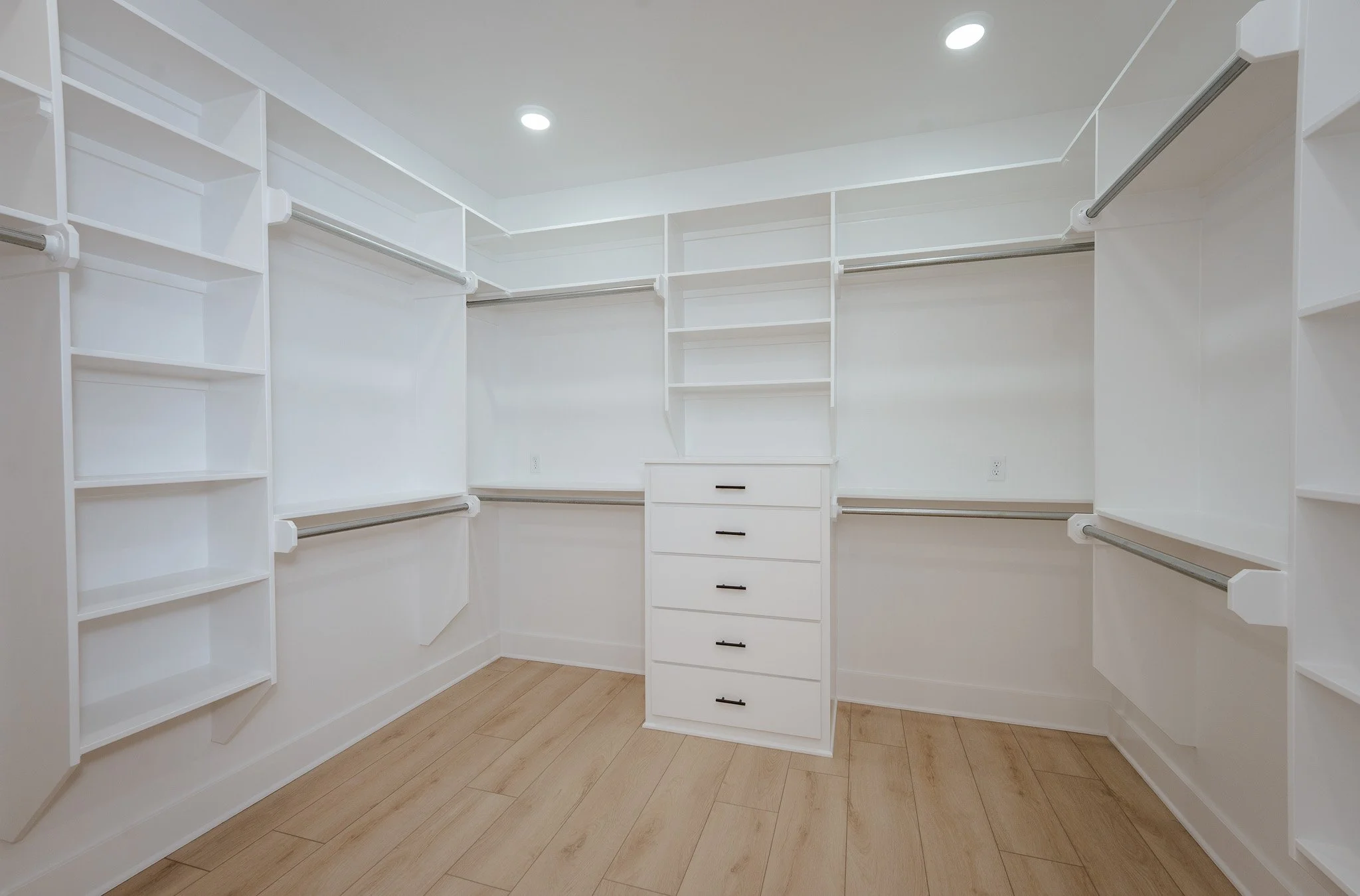 Empty walk-in closet with white shelving, drawers, and hanging rods on all sides, and light-colored wooden flooring.