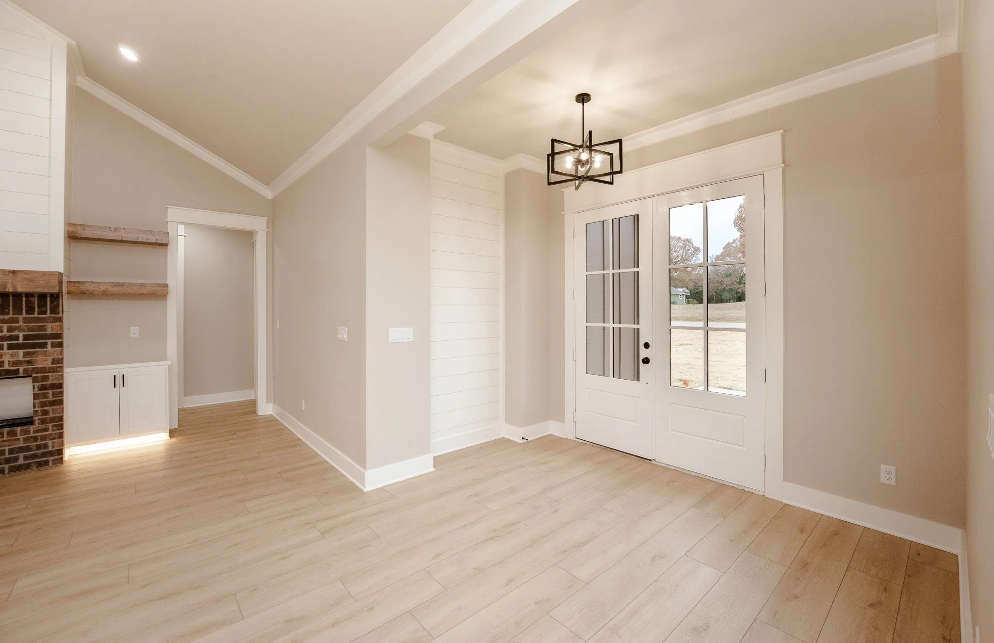 Empty living room with beige walls, light wood flooring, white trim, a white door with glass panels, and a black chandelier hanging from the ceiling.