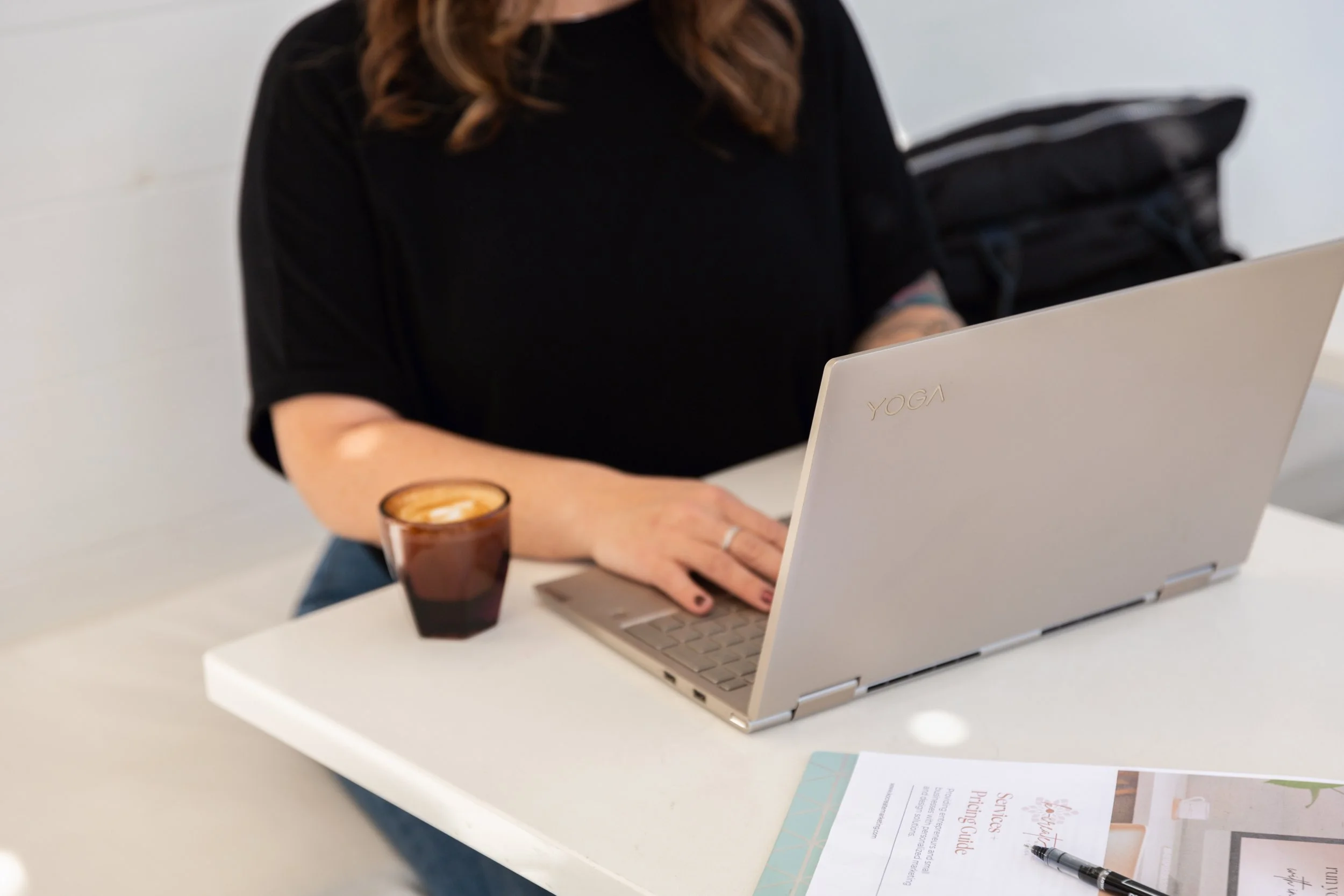 Woman working on a laptop at a minimalist white desk with coffee and marketing materials