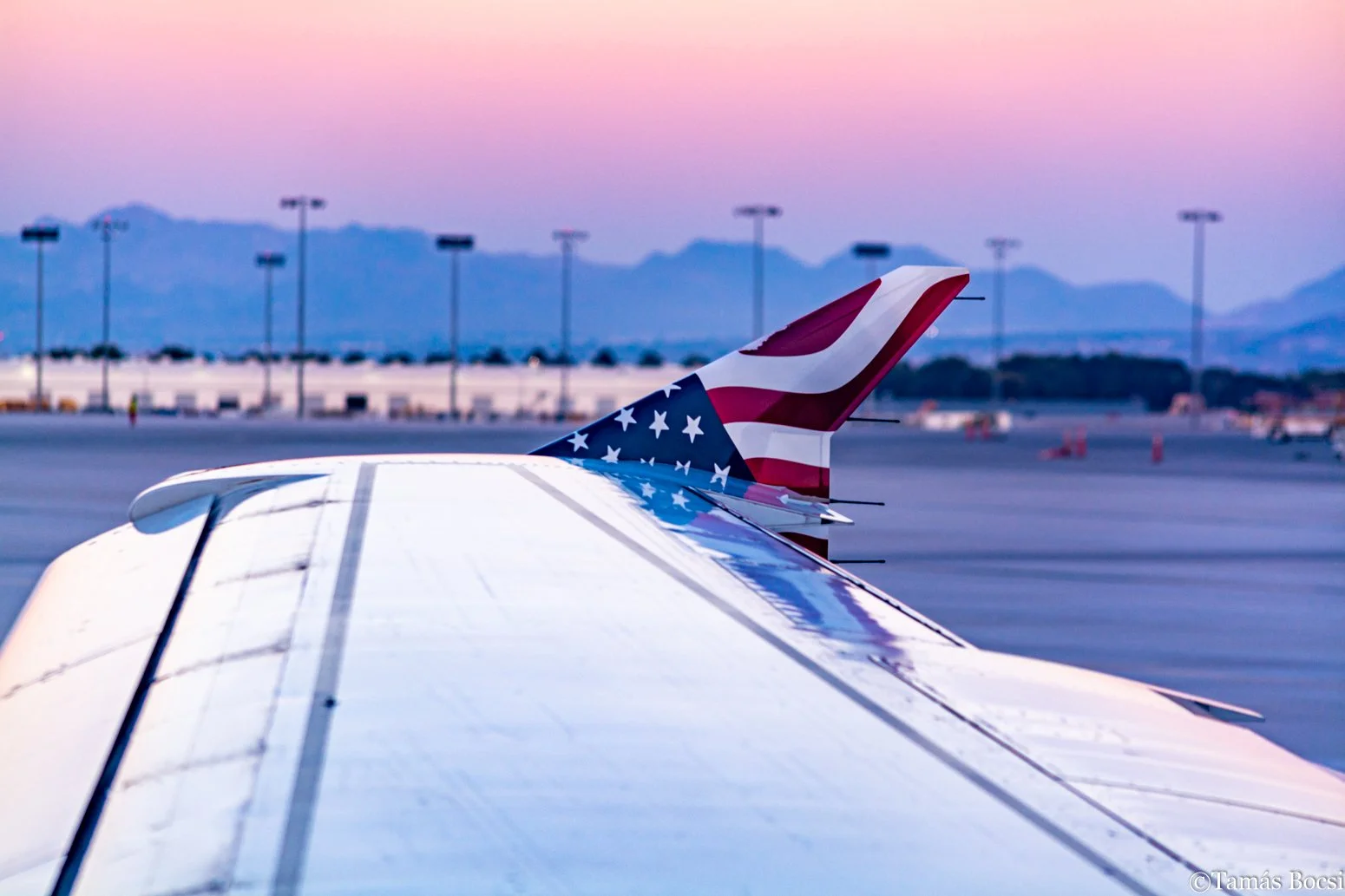 Close-up of the tail of an airplane painted with the American flag, on an airport tarmac at sunset or dawn with mountains in the background.
