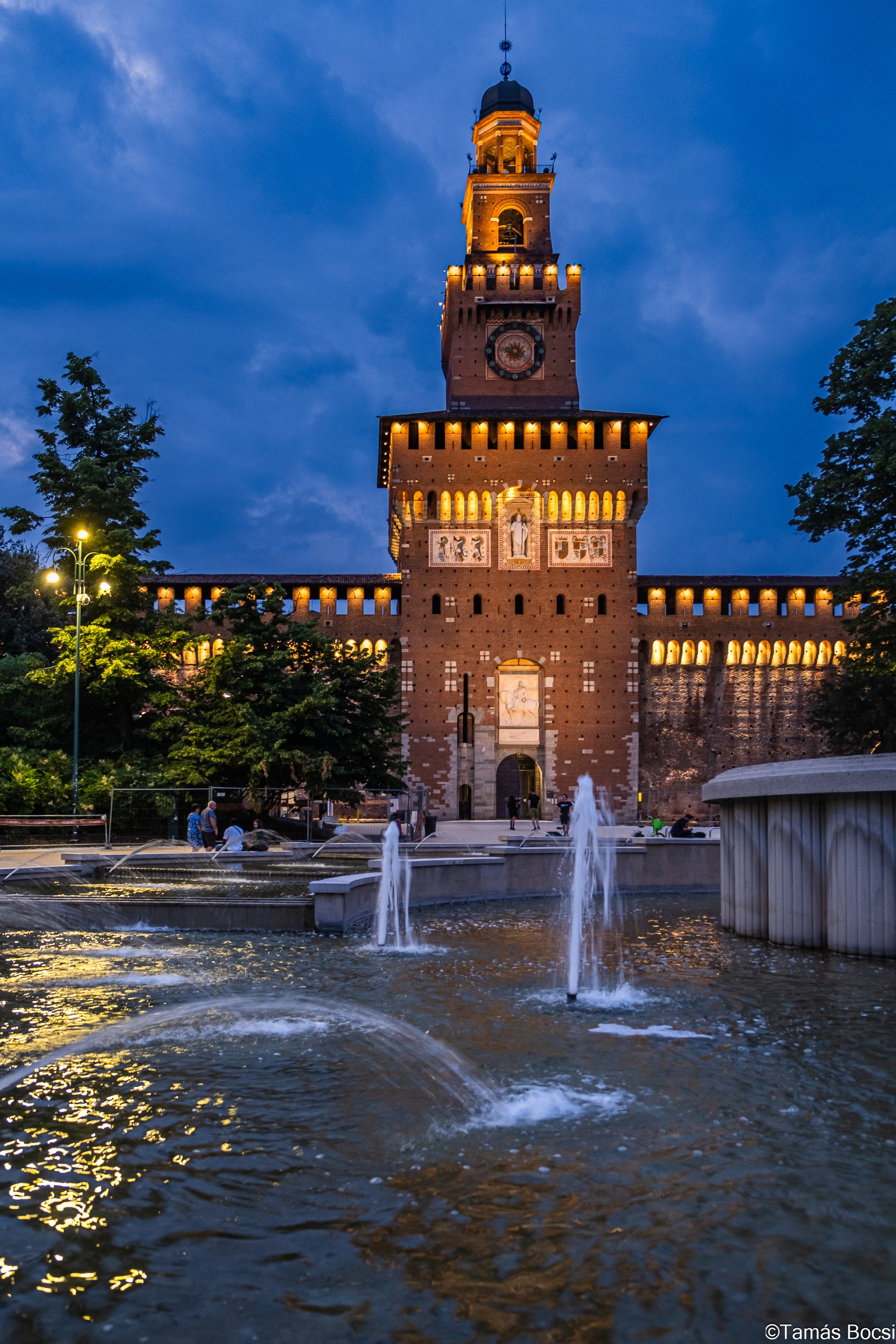 Nighttime view of a historic brick castle with a tall tower, illuminated with warm lights, and water fountains in the foreground.