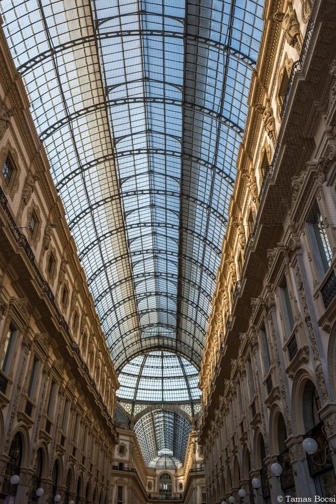 Interior view of a grand shopping arcade with a high glass arched ceiling and ornate classical architecture on either side.