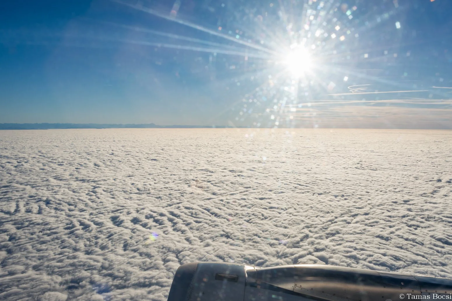 View from an airplane window showing a vast snowy landscape with the sun shining brightly in a clear blue sky.