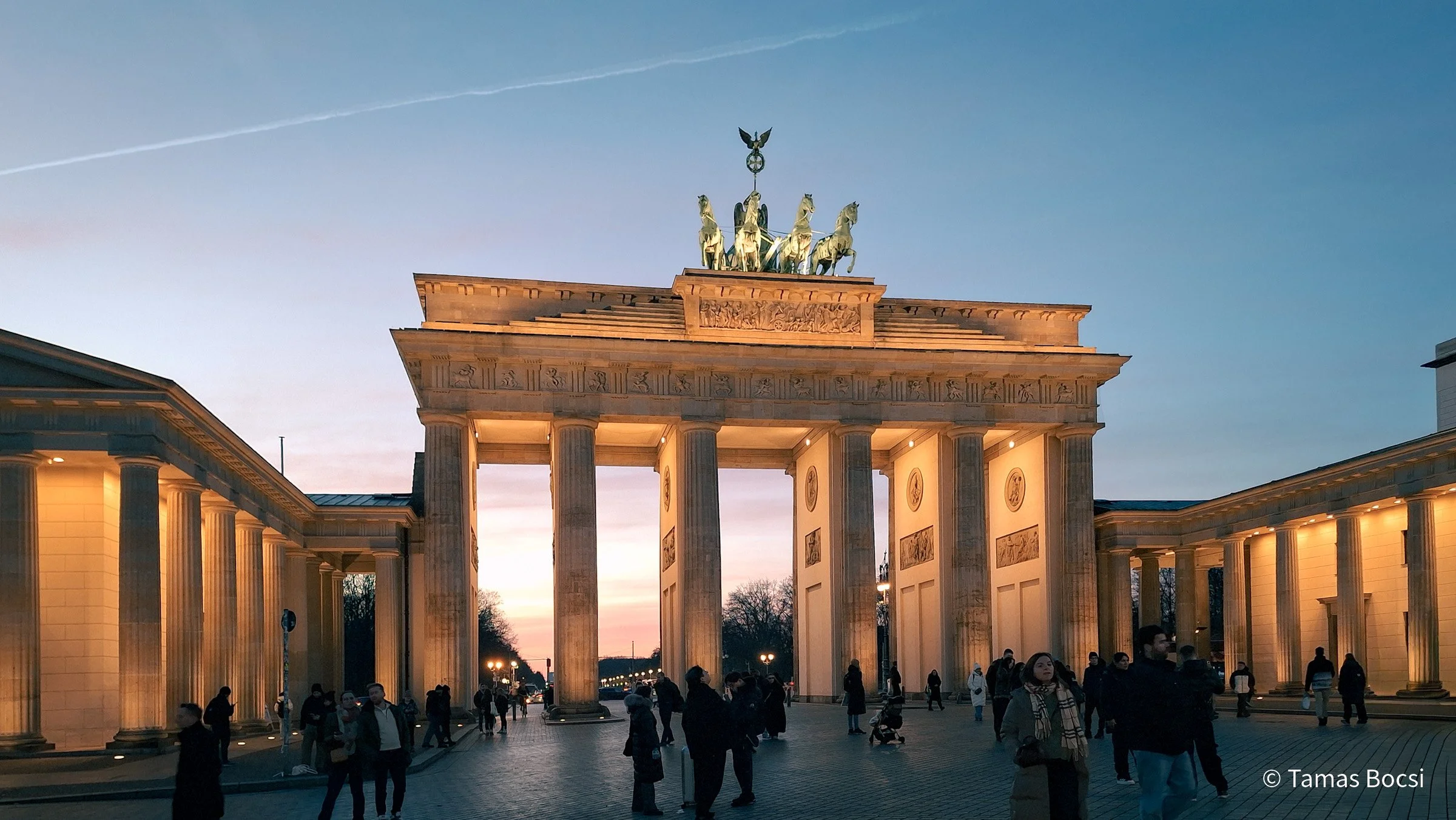 Nighttime view of the Brandenburg Gate in Berlin, Germany, with illuminated columns and a clear sky, people walking and standing around.