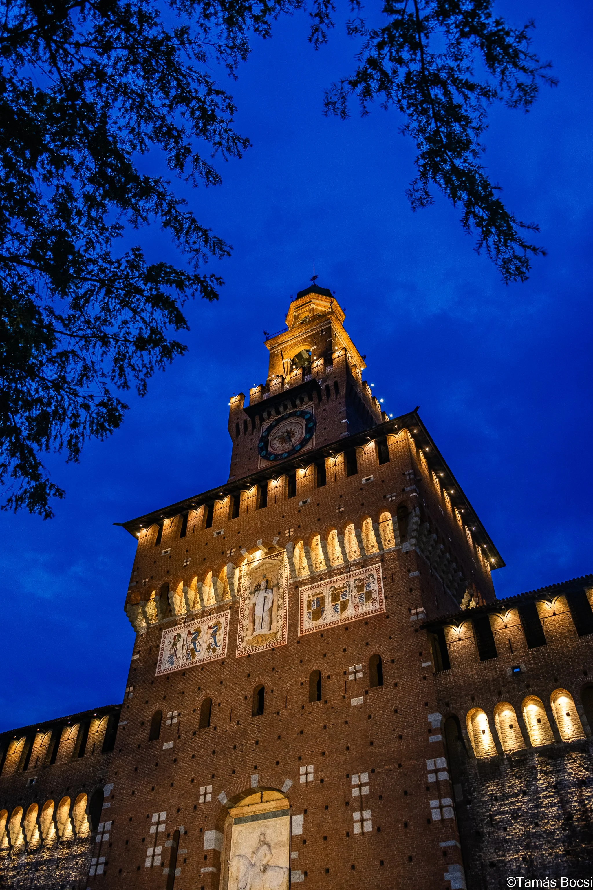 A tall, illuminated historic brick tower with decorative murals and a clock, viewed from below during twilight, with tree branches framing the top of the image.
