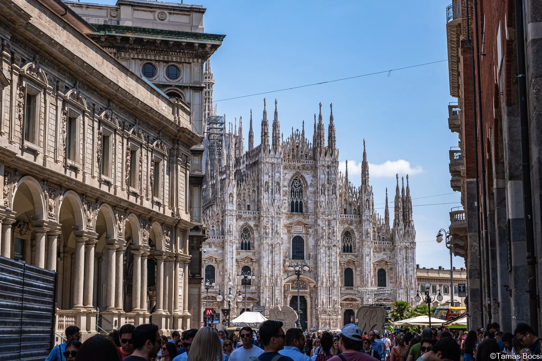 Crowd of people walking in a busy city street with the Milan Cathedral in the background.