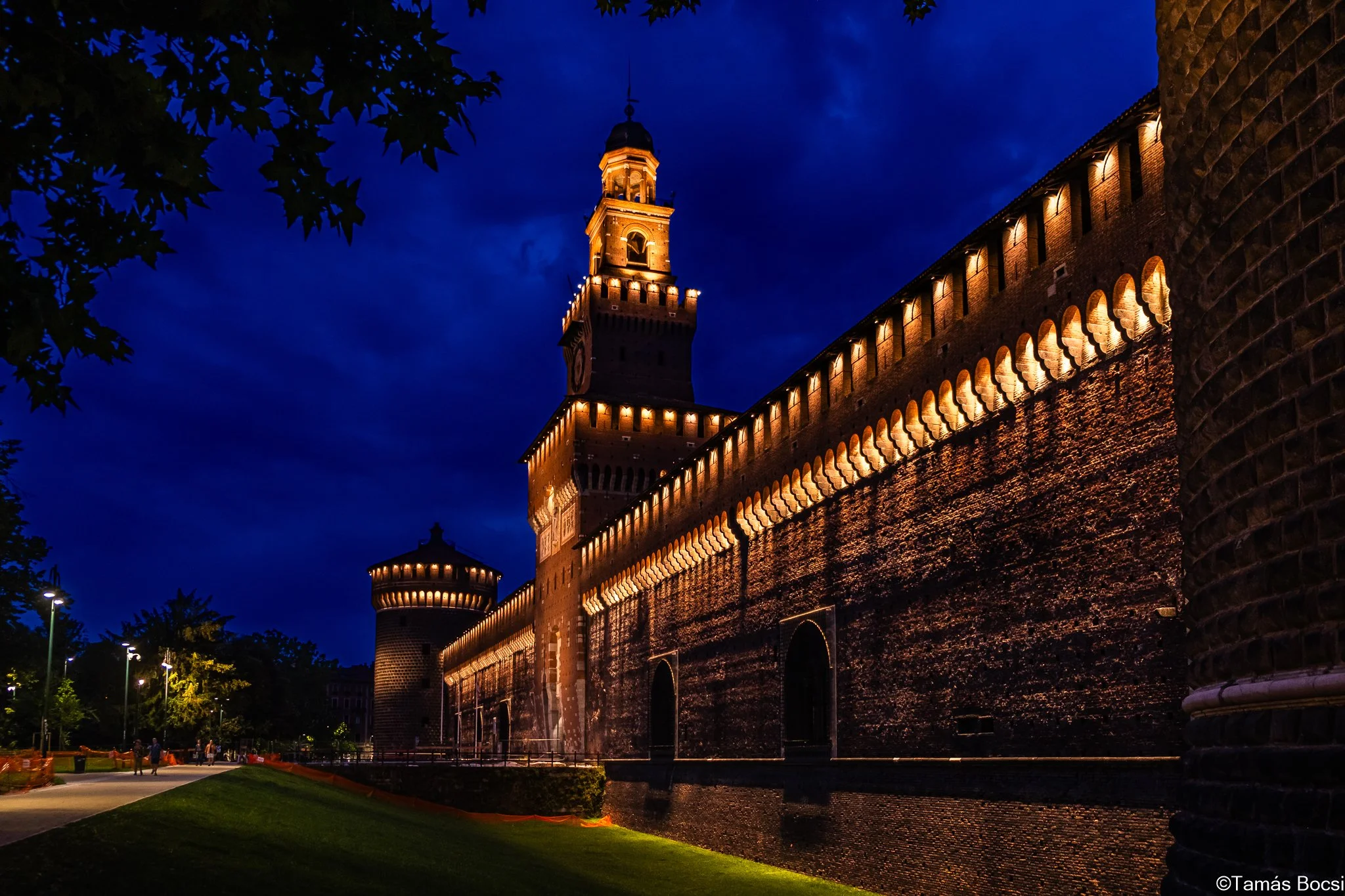 A historic brick fortress illuminated at night with tall towers and bright lights, set against a dark blue sky.