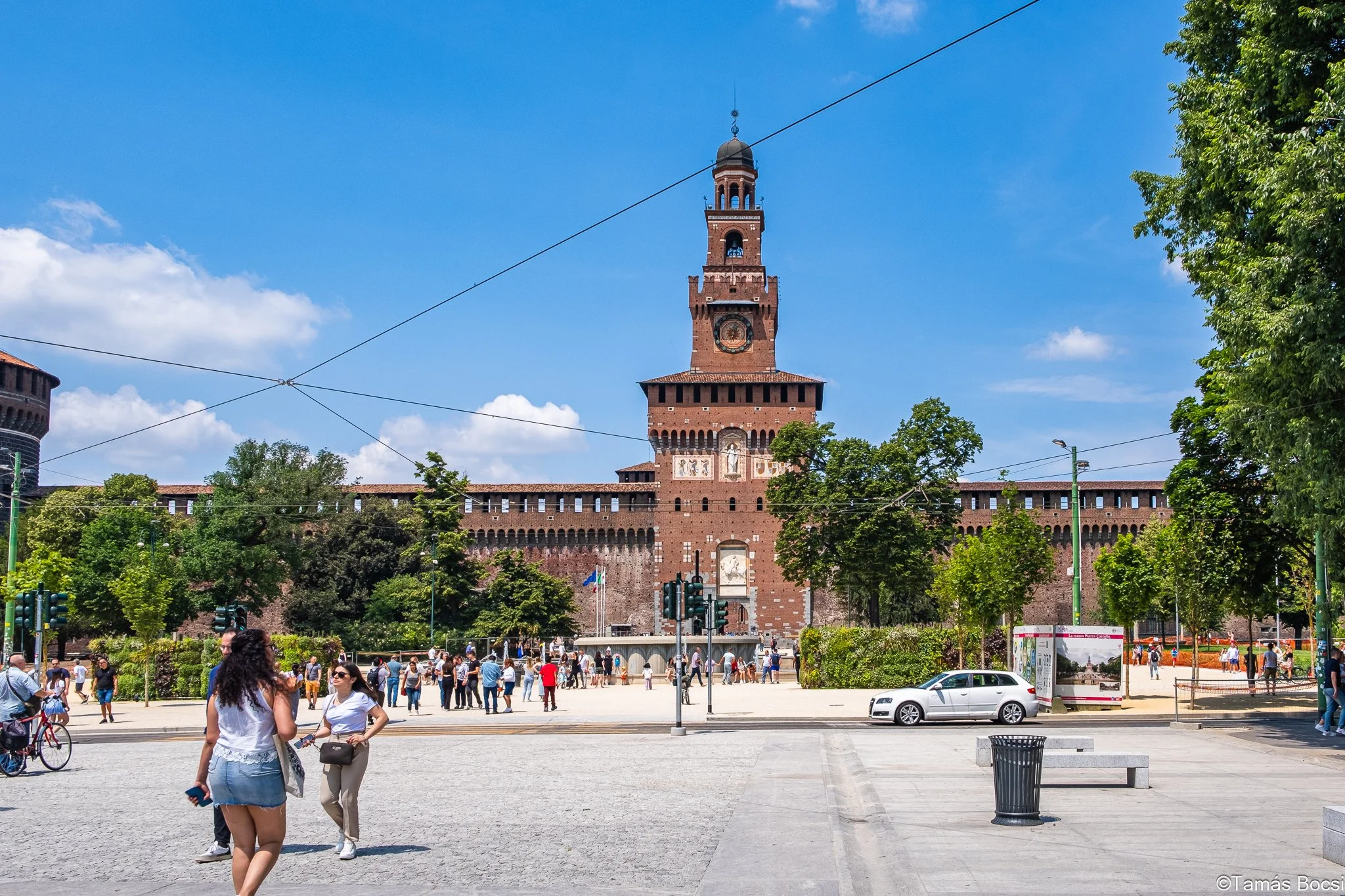 People walking and gathering in front of a large historic brick castle with a tall clock tower, surrounded by trees, on a sunny day with a blue sky.