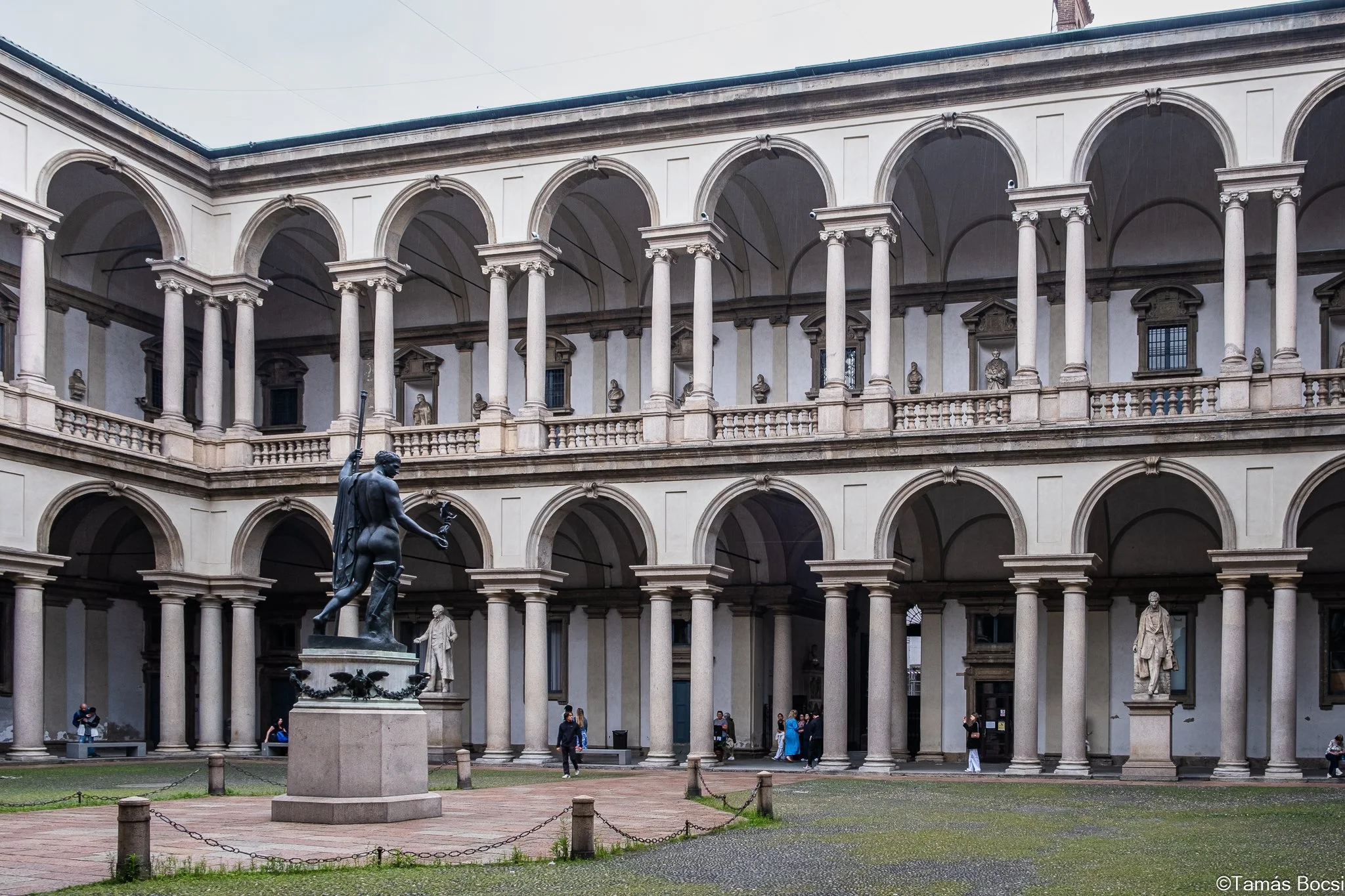 A historic courtyard with an ornate two-story classical building featuring arches and columns, and a statue of a woman holding a sword and a shield in the center.