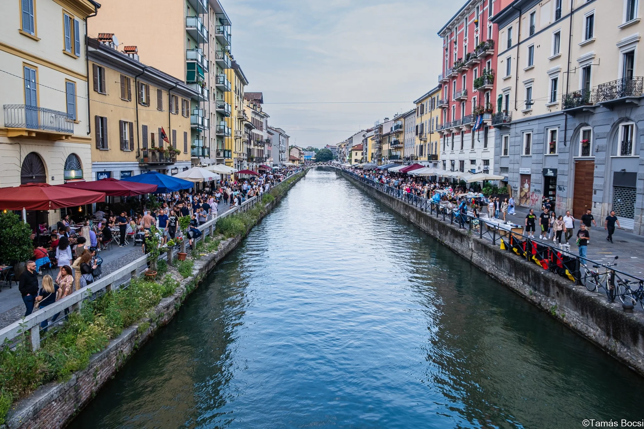 A canal lined with colorful buildings and crowded outdoor cafes on both sides, with people walking and dining along the waterway.
