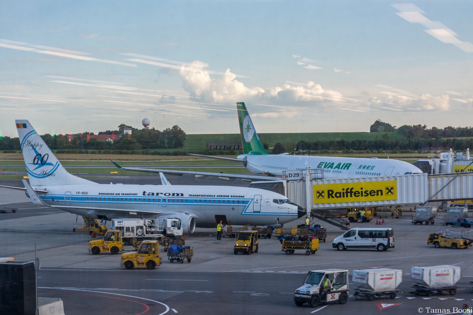 Two airplanes parked at the gate of an airport, one Romania Air Transport and one EVA Air, with various ground service vehicles and personnel around them.