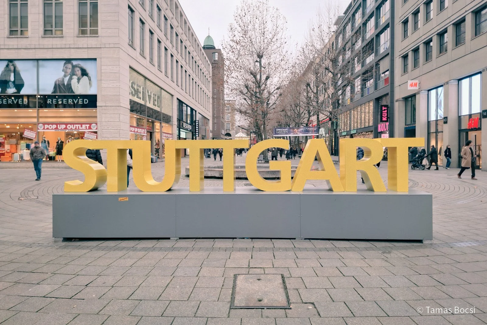 Large yellow letters spelling 'STUTTGART' on a grey platform in a city street with shops and people walking.