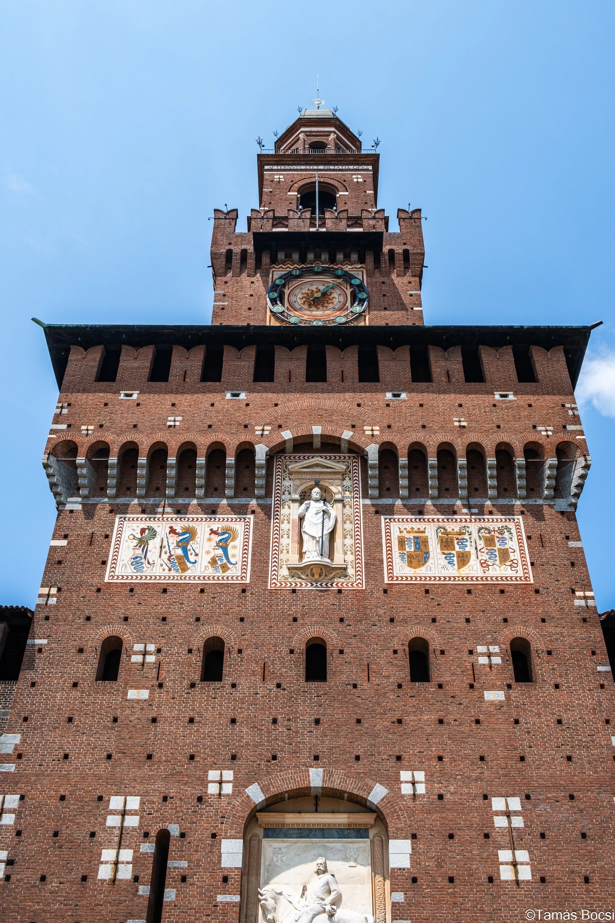 A tall brick clock tower with architectural details, a large clock face, and decorative murals, set against a blue sky.