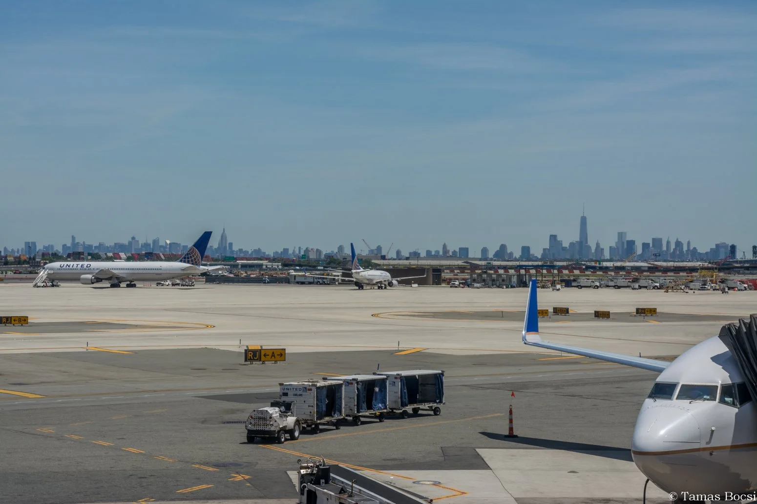 View of an airport tarmac with airplanes, ground vehicles, and a city skyline in the background, including the Empire State Building and One World Trade Center.