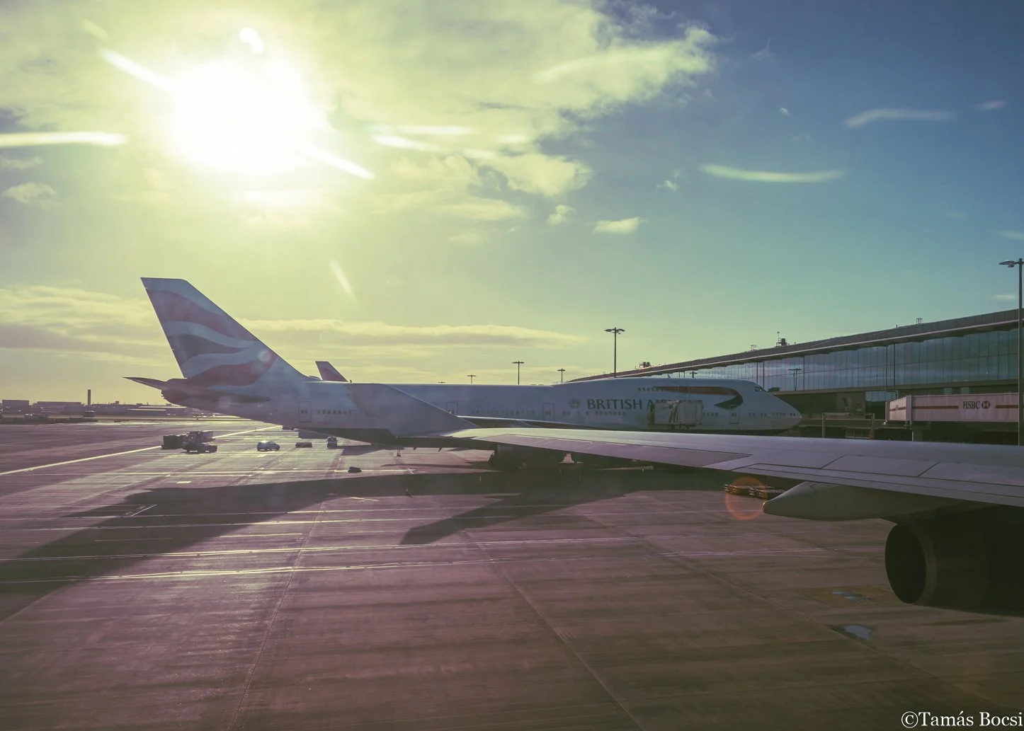 View of an airplane parked at an airport tarmac during sunset, with the sun shining bright in the sky and a large glass airport terminal in the background.