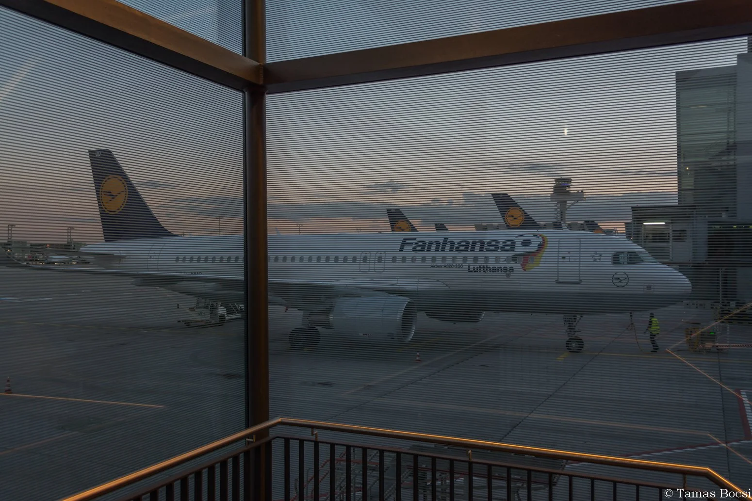 View of Airbus airplane at airport gate through glass window with horizontal lines, during dusk, with airport infrastructure and personnel in background.