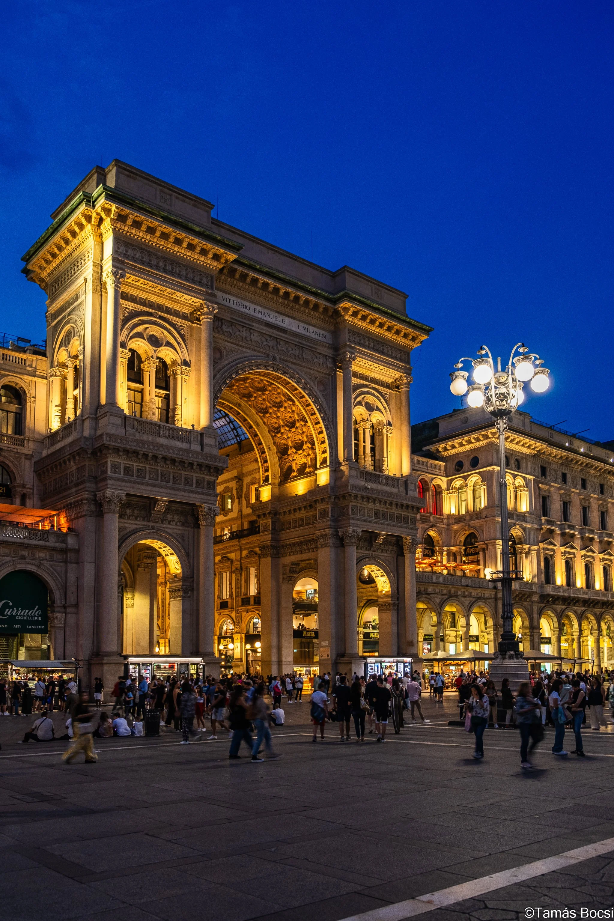Nighttime view of Galleria Vittorio Emanuele II in Milan, Italy, with lit-up architecture and crowds in the square.