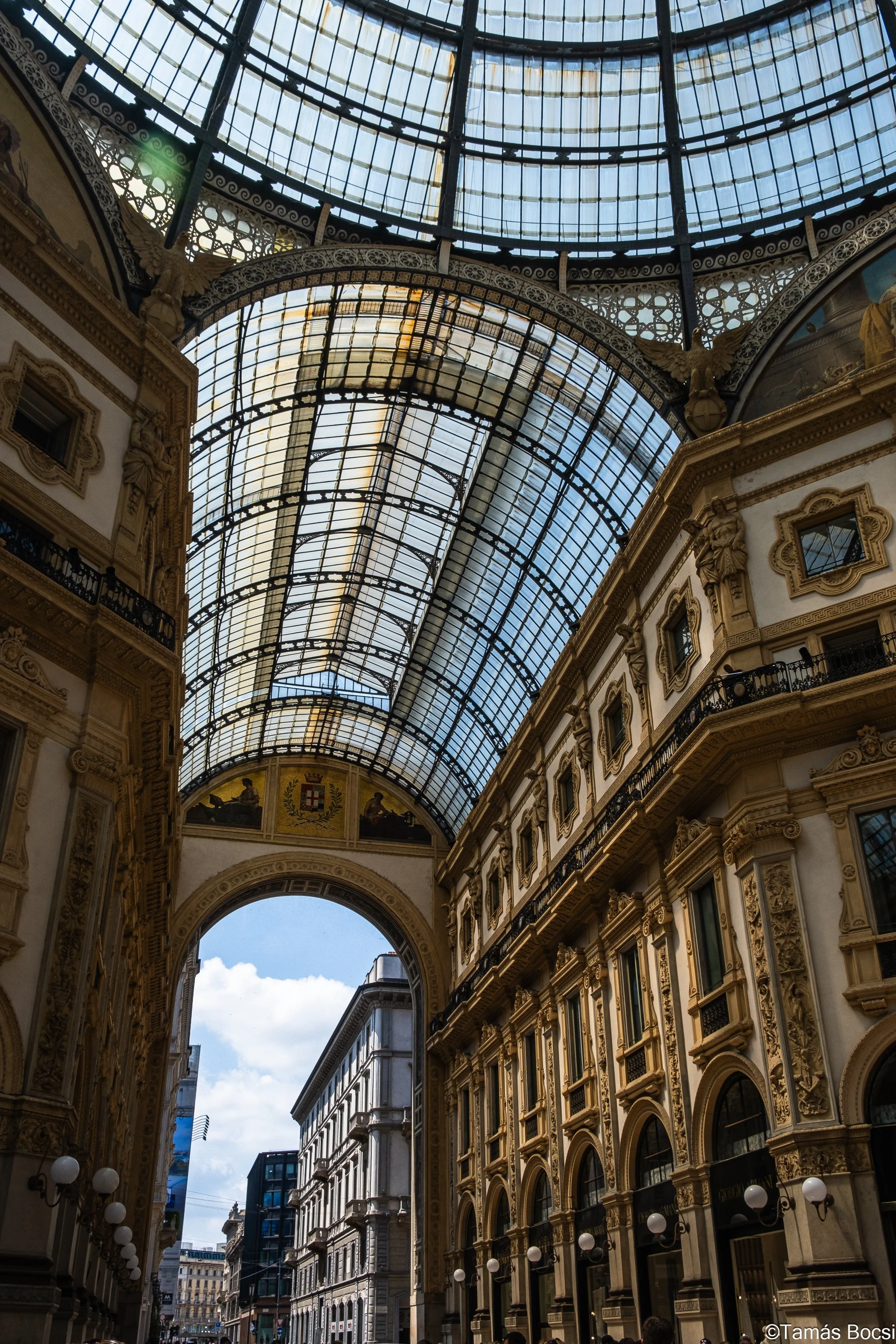 Interior view of a grand historic building with ornate gold and white detailing, large arched windows, and a glass domed ceiling with lattice framework.