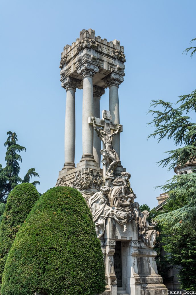 A tall stone monument featuring a crucifix with Jesus Christ on the cross, surrounded by detailed sculptures of multiple figures, and topped with four pillars supporting a decorated platform.