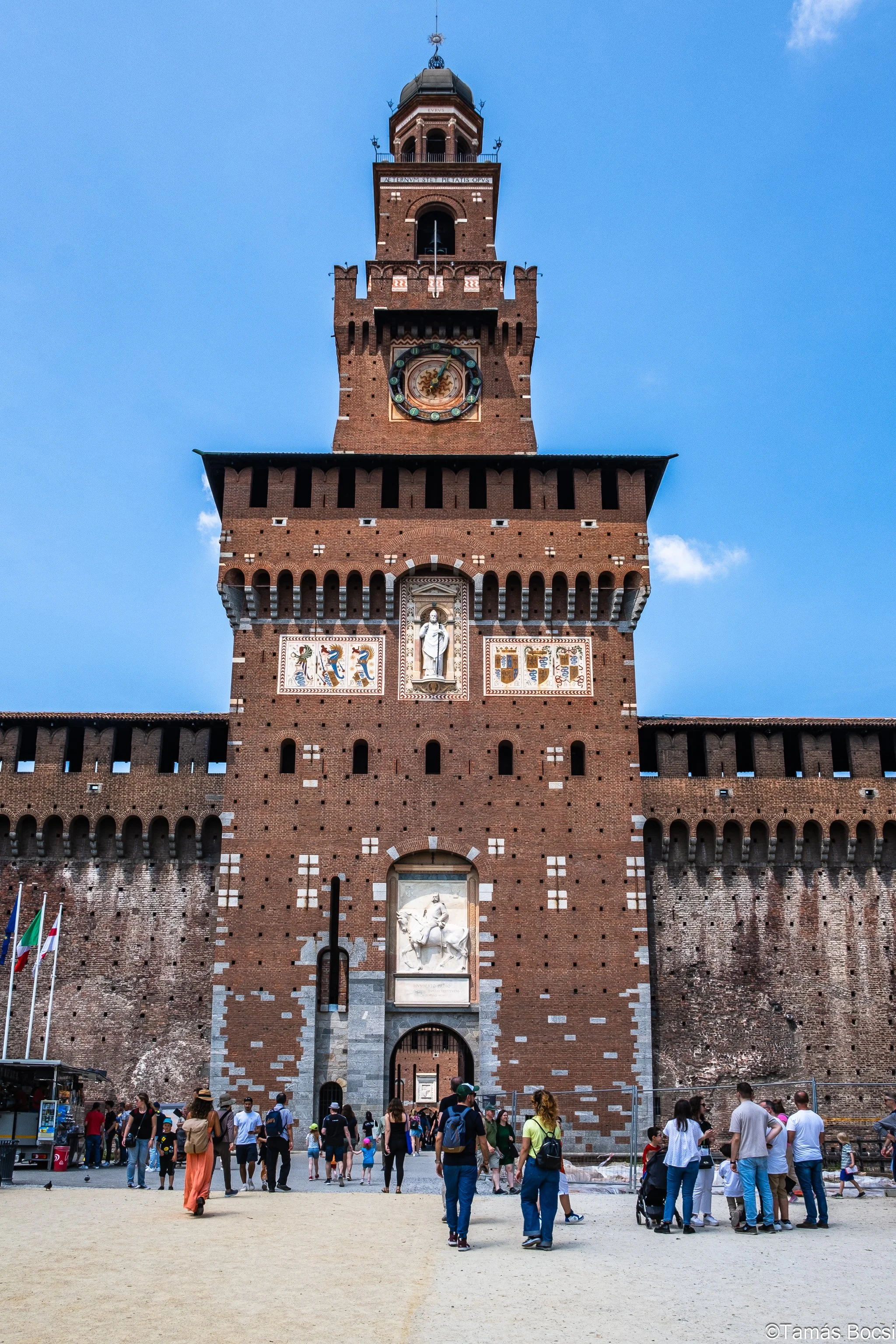 A tall brick medieval castle tower with a clock and statues, crowds of people at the base, clear blue sky in the background.