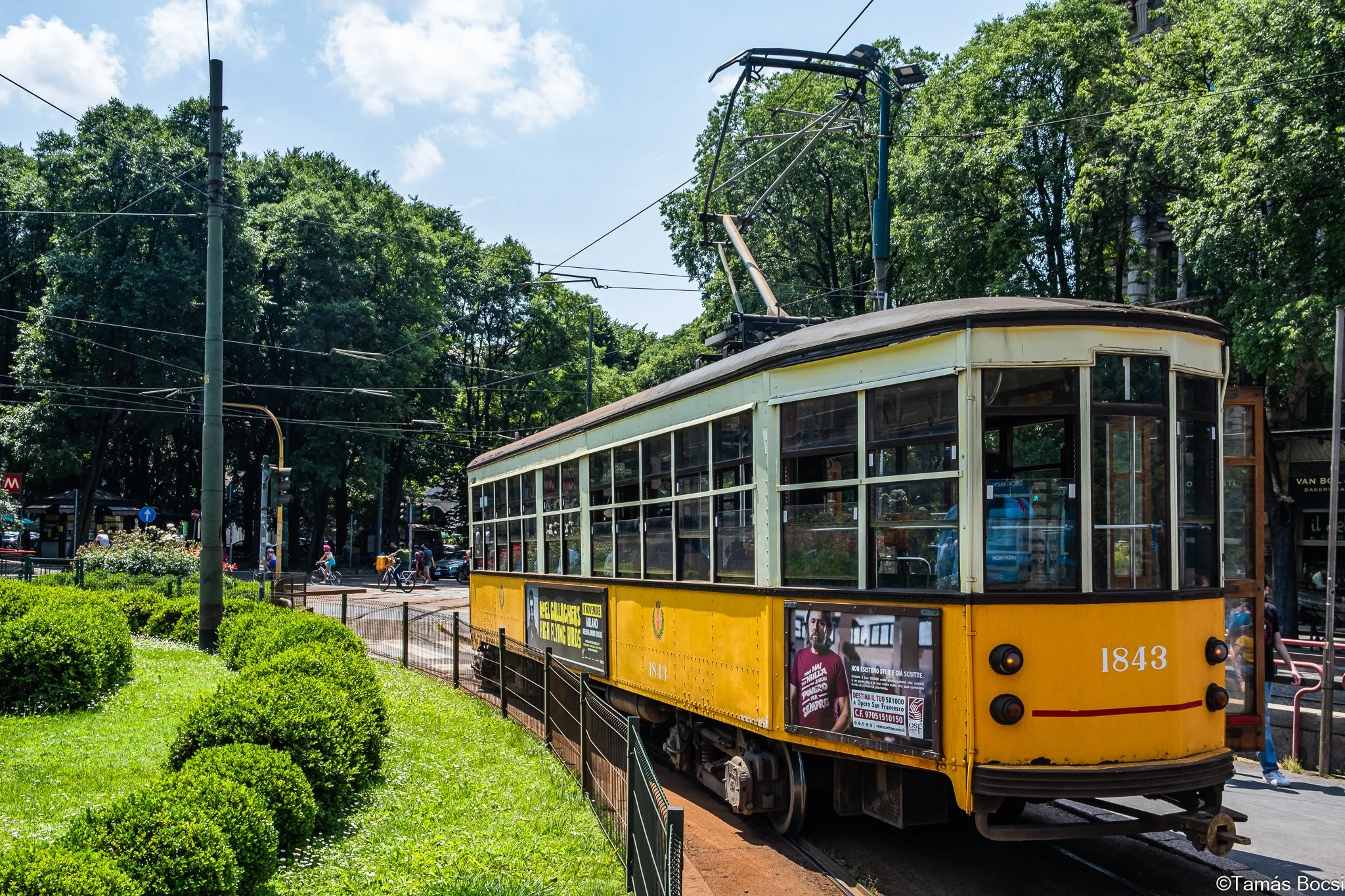 Vintage yellow tram traveling on a city street with trees and a park on side, people walking and riding bikes in the background.