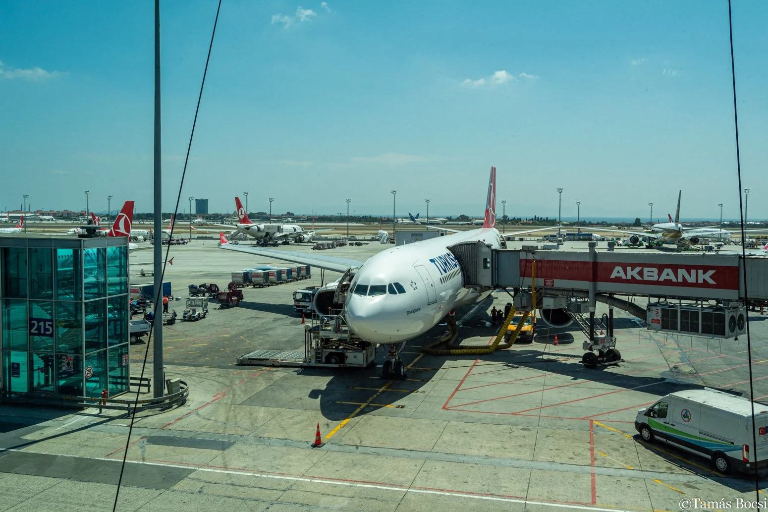 View of an airport tarmac with a Turkish Airlines airplane connected to a passenger gate, surrounded by airport vehicles and several other airplanes parked in the background under a blue sky.