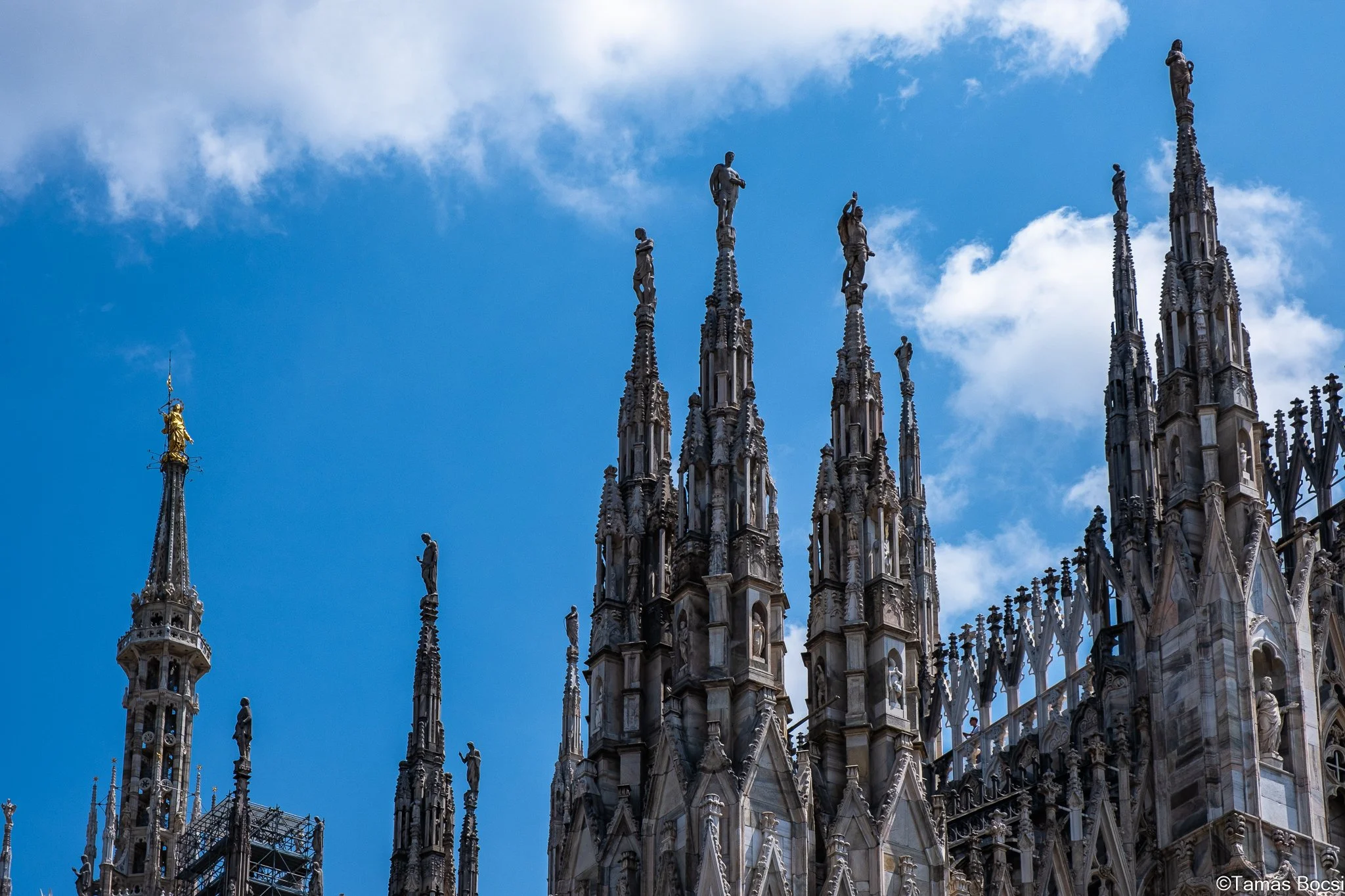 Close-up of the intricate Gothic spires and statues on the roof of a cathedral, with a blue sky and clouds in the background.