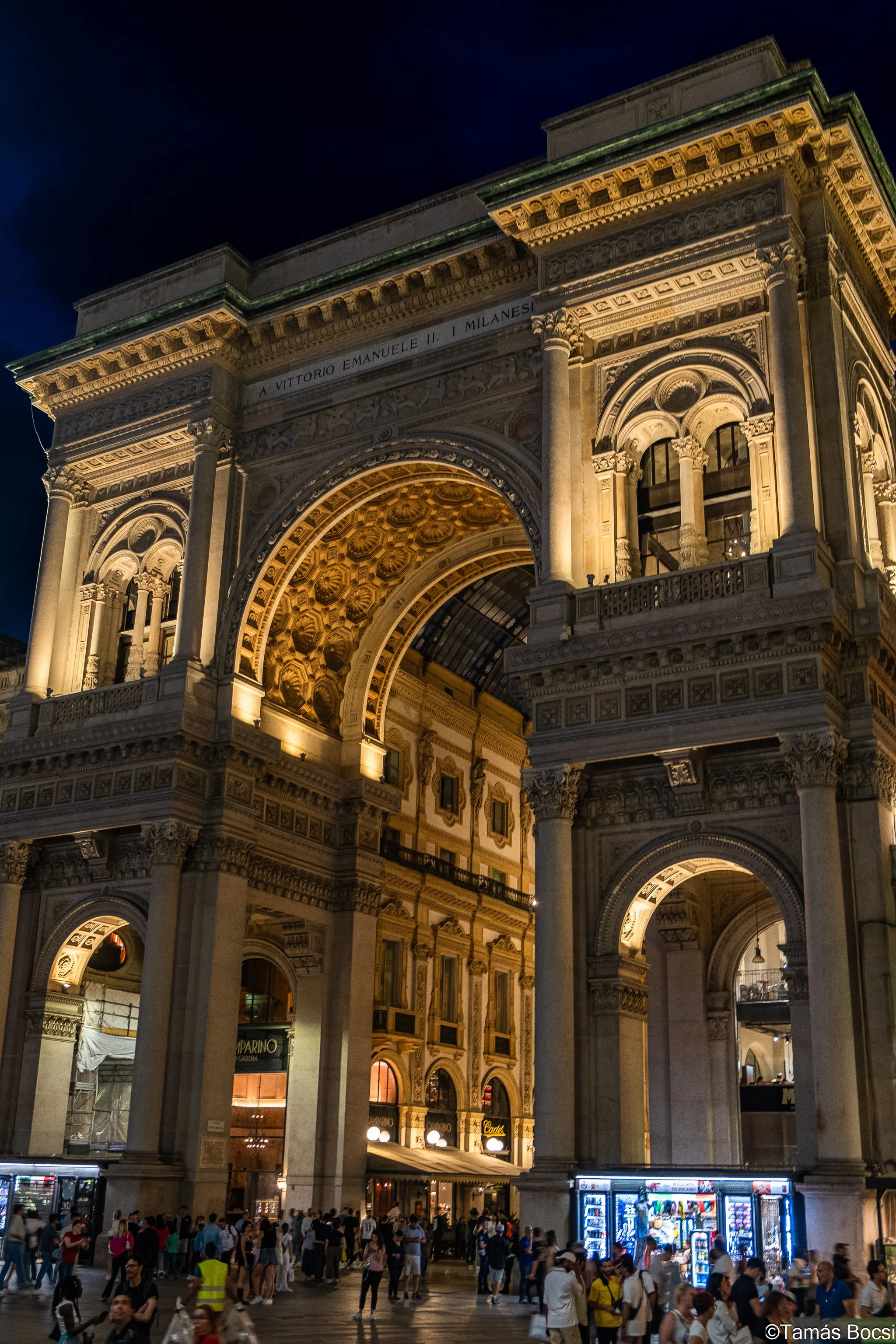 Night view of the Arch of Triumph in Milan, Italy, with many people gathered at the base, illuminated by lights.