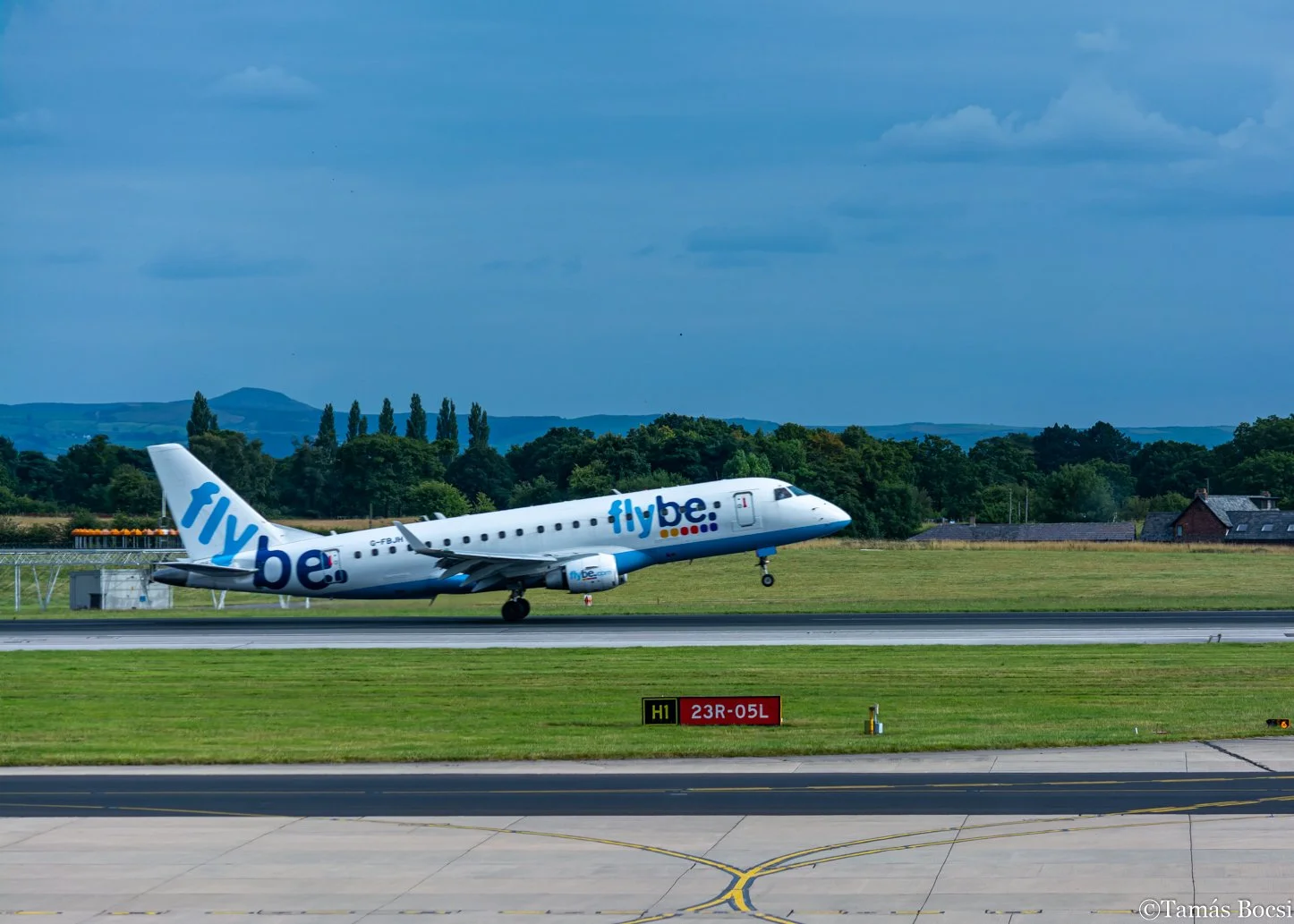 A Flybe commercial airplane taking off from an airport runway with a background of trees and hills under a partly cloudy sky.