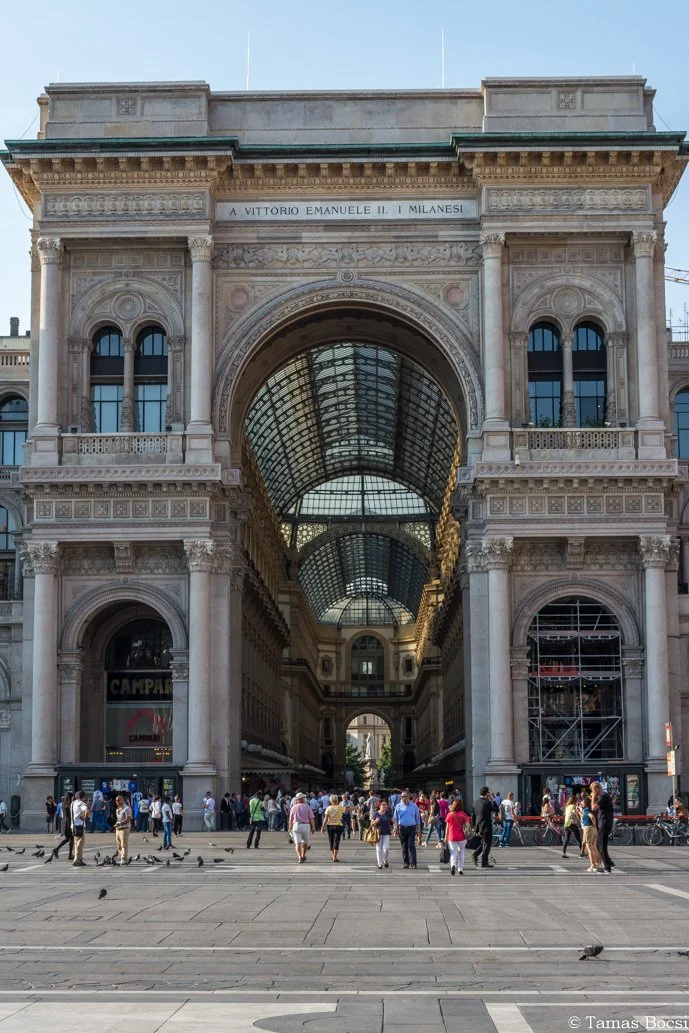 A historic building with ornate architecture featuring a large arched opening in the center, crowned with a glass-covered passageway, and a crowd of people walking in front.