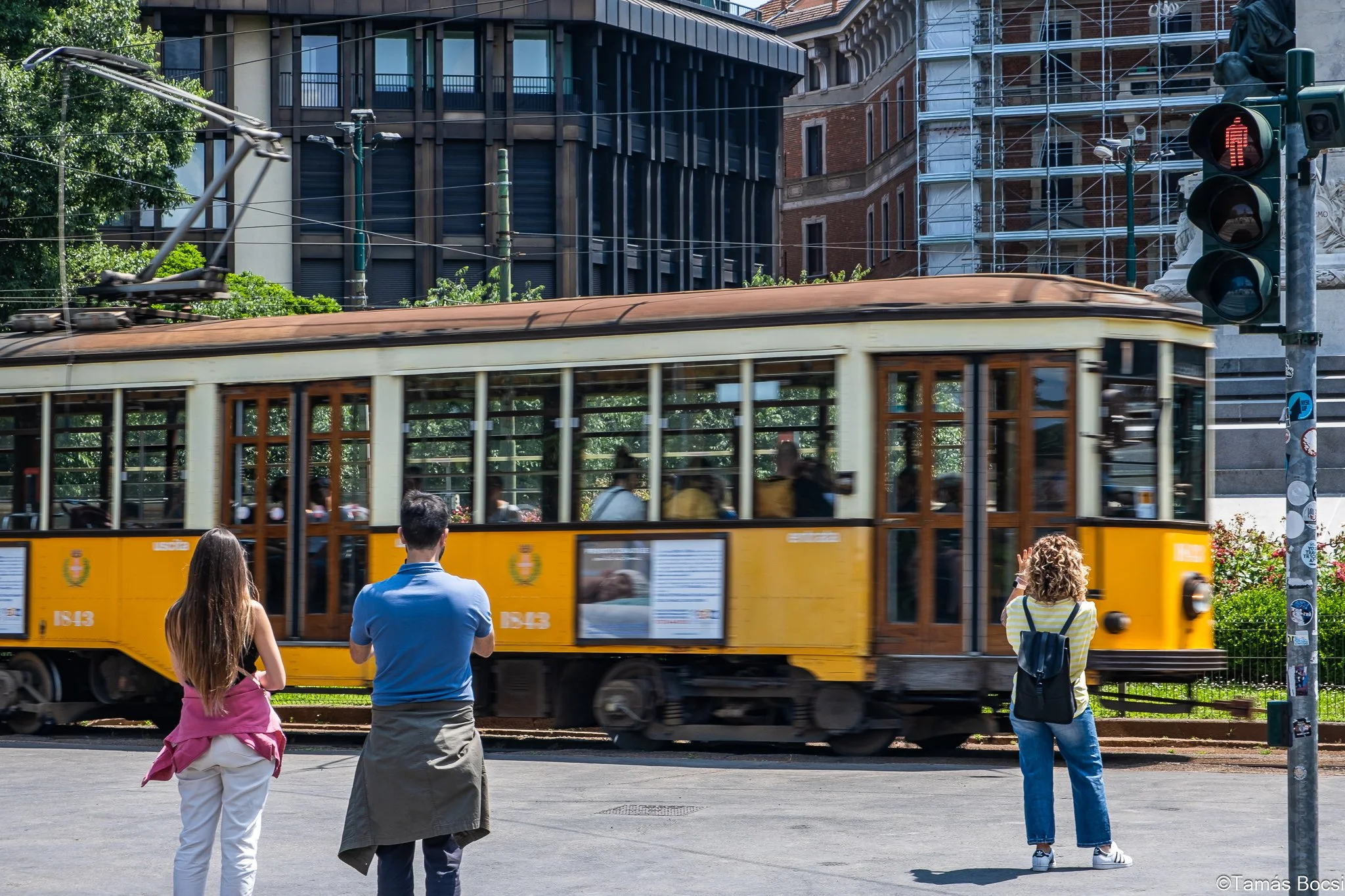 Three people waiting to cross the street as a yellow tram passes by in an urban area with tall buildings and traffic lights.