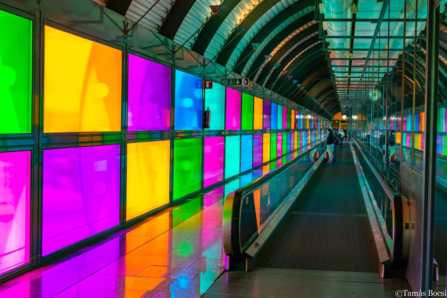 An airport moving walkway featured through a corridor with glass walls on one side displaying colorful illuminated panels in green, yellow, pink, purple, and blue. Passengers are visible in the distance carrying luggage.