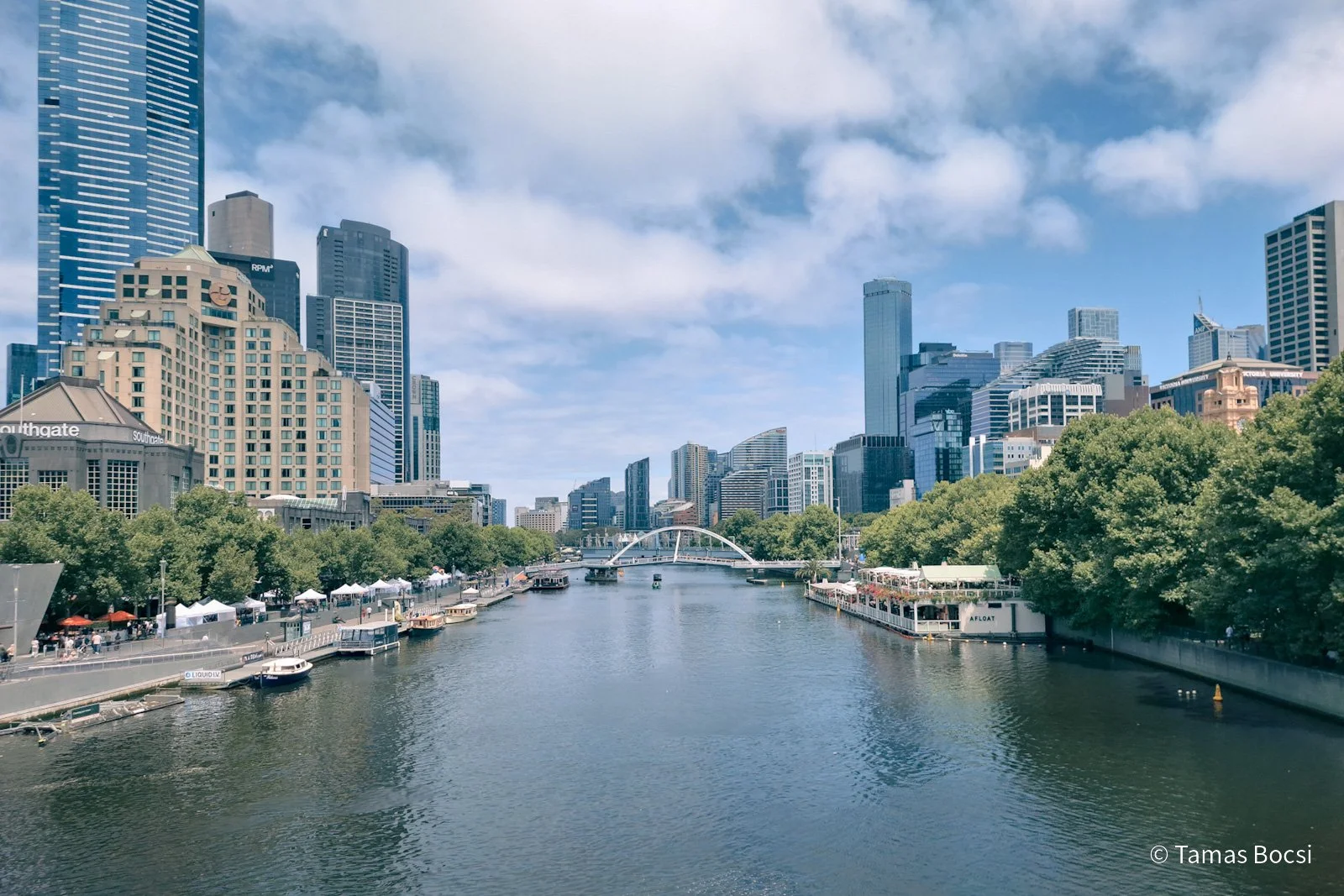 City skyline with tall modern buildings, a river with boats, trees lining the riverbanks, and a footbridge in the distance on a bright, partly cloudy day.