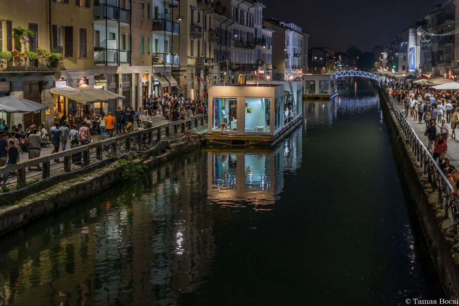 Nighttime scene of a busy canal street with people walking along the sidewalks, crowded outdoor cafes, and a modern floating bar or restaurant on the water, with illuminated buildings and a small bridge in the distance.